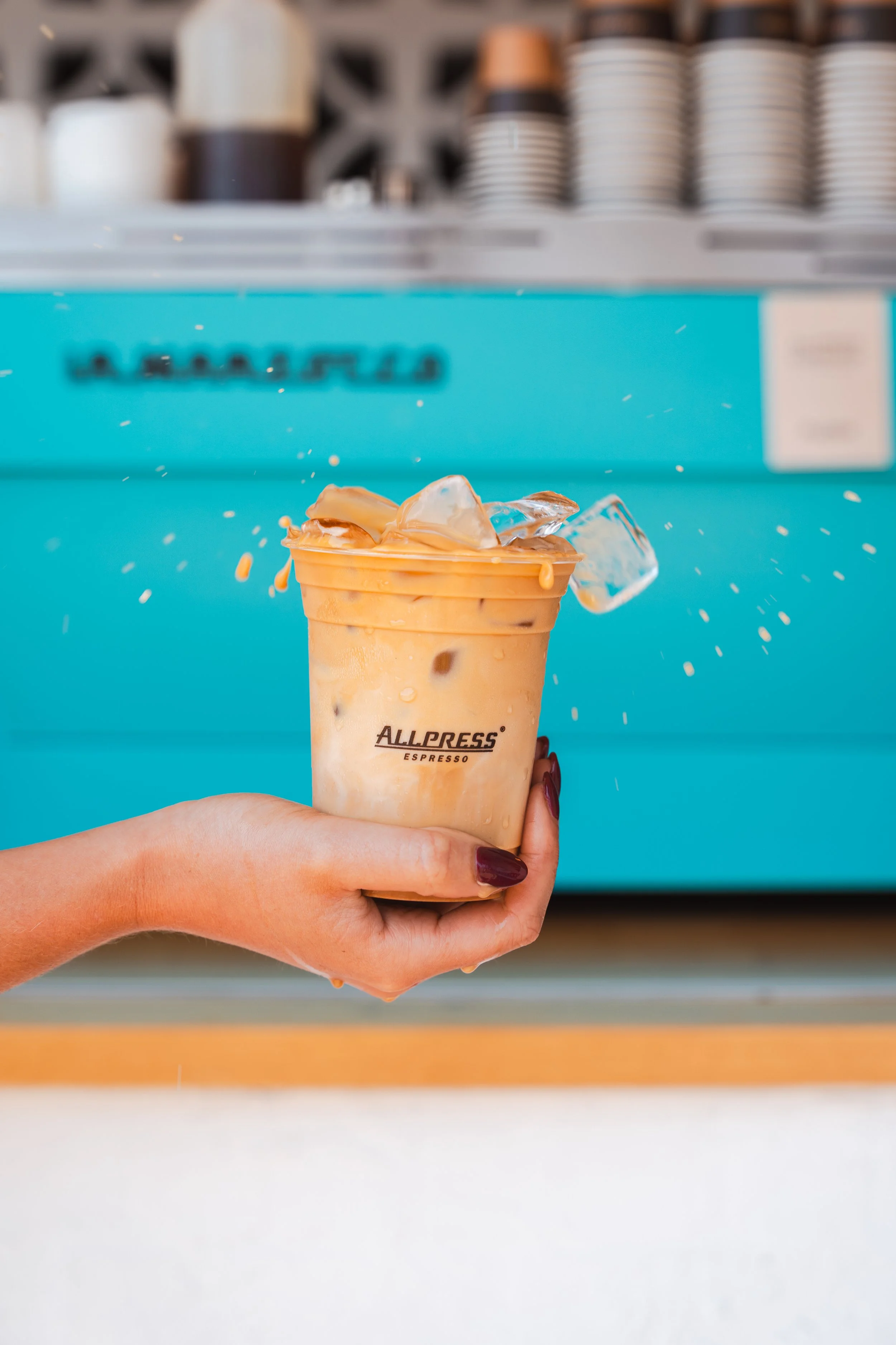 Hand holding a plastic cup of iced coffee with ice cubes, with a bright blue food truck in the background.