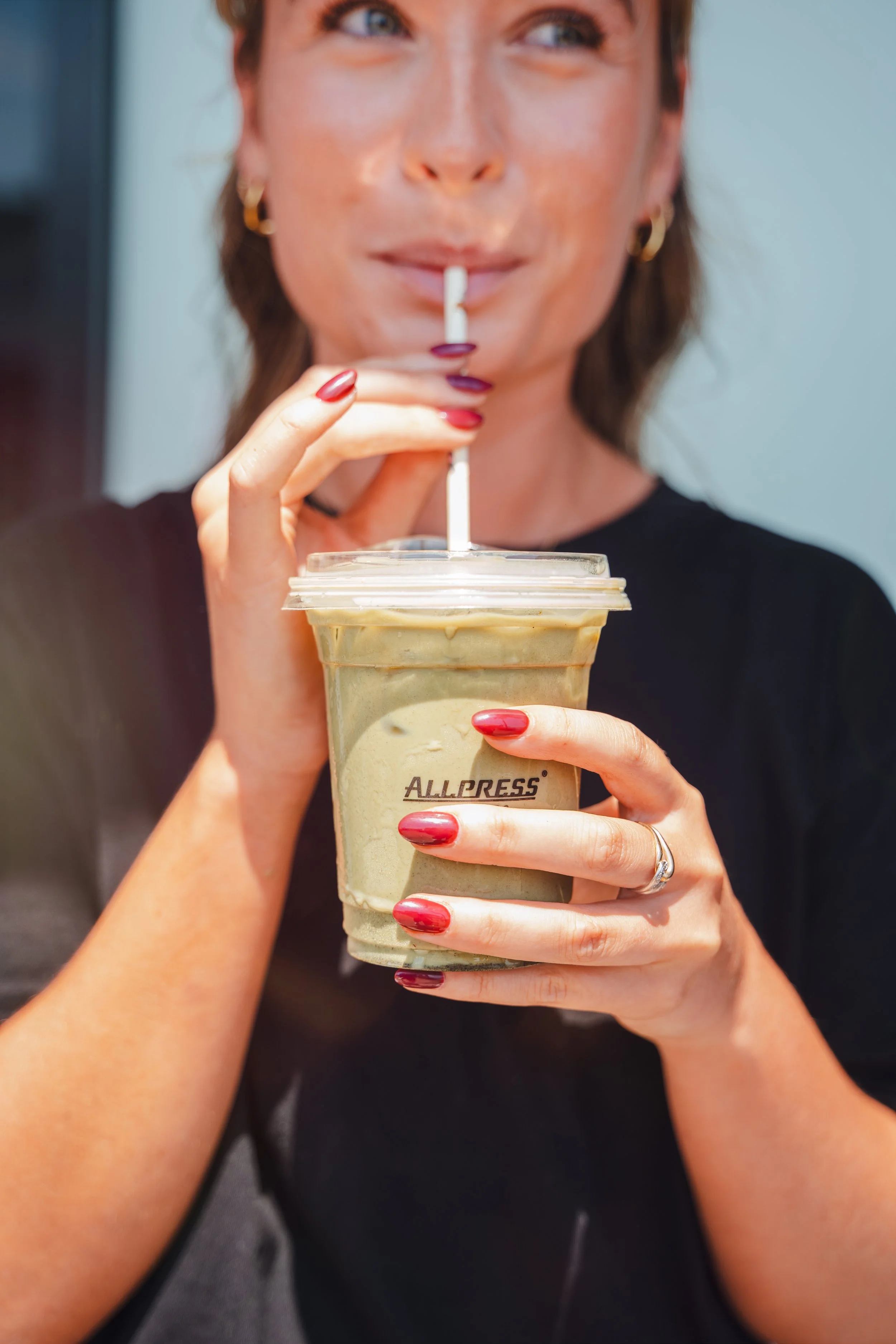 Woman with painted nails enjoying iced coffee or matcha latte, holding a straw in a clear plastic cup labeled 'Allpress'.