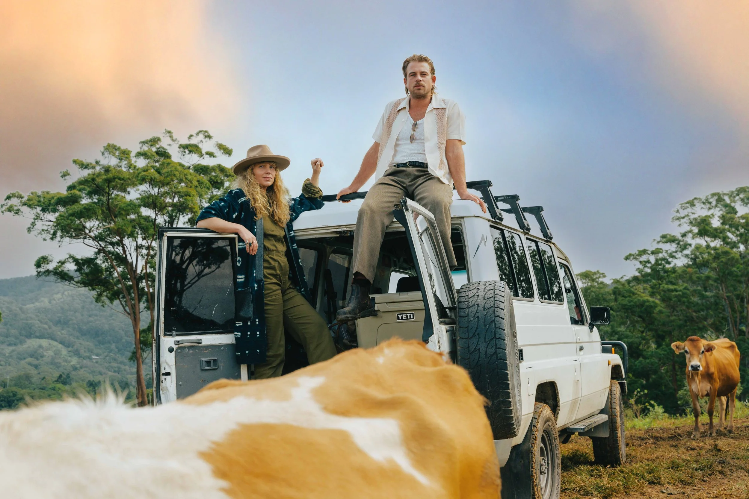 A man and woman on top of a white off-road vehicle with dogs nearby in a rural setting with trees and hills in the background.