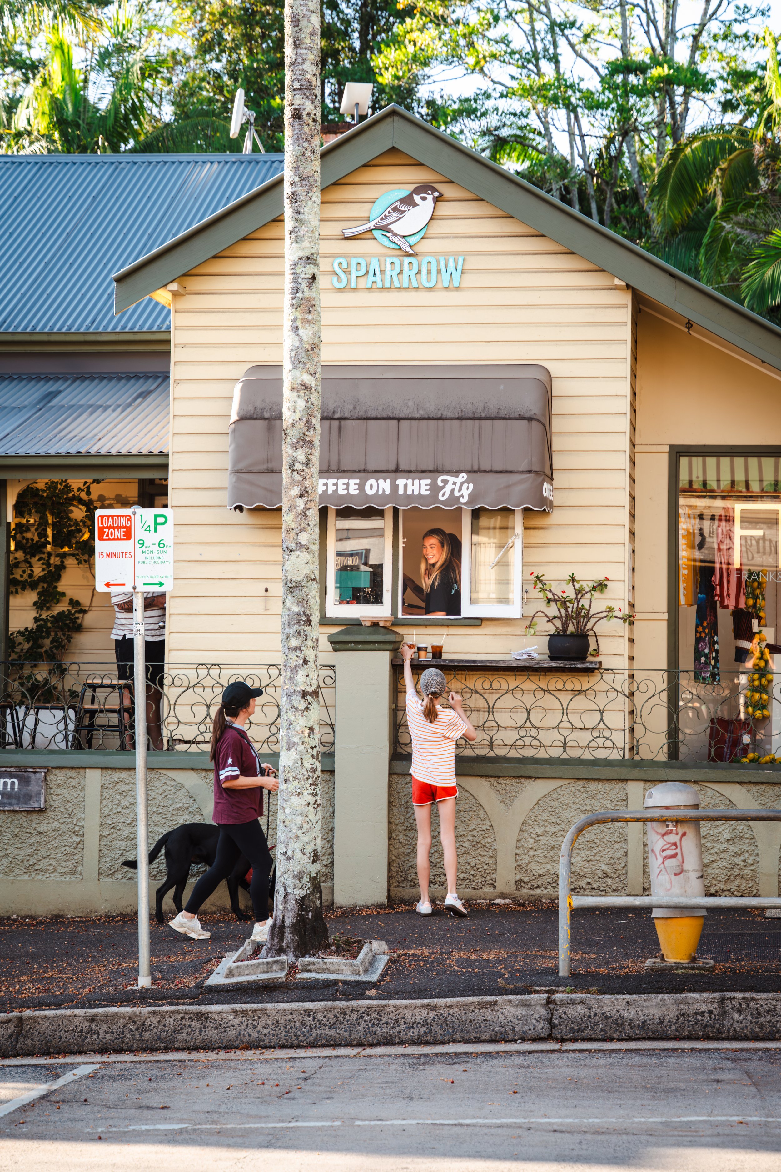 A small building with a sign that says 'Sparrow' and a bird illustration, with a coffee stand underneath and people standing outside, including a woman in a striped shirt, red shorts, and a woman with a dog walking by.