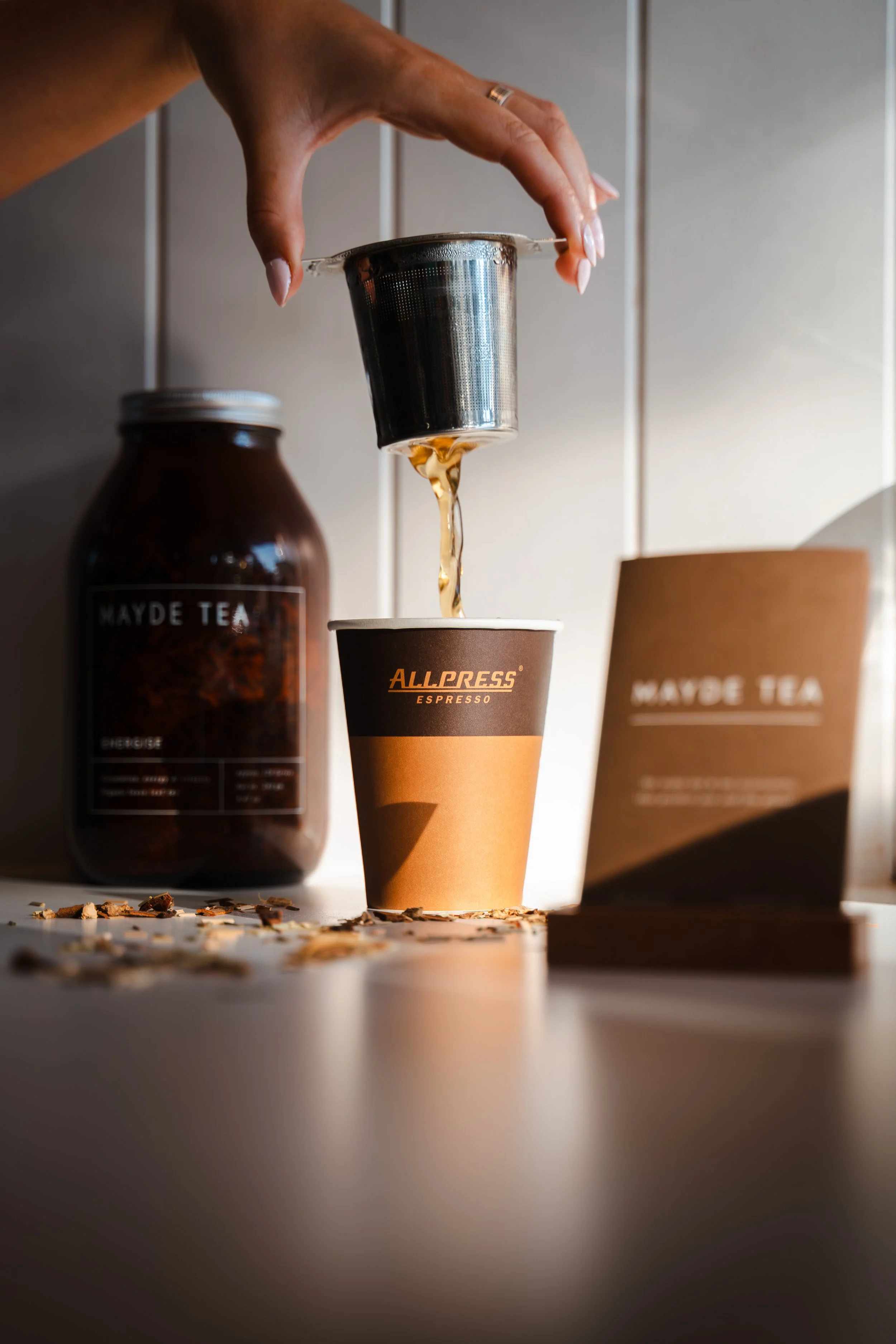 A hand holding a stainless steel tea infuser above a paper cup labeled 'ALLPRESS ESPRESSO', with spilled tea leaves and a jar of tea labeled 'MAYDE TEA' in the background.