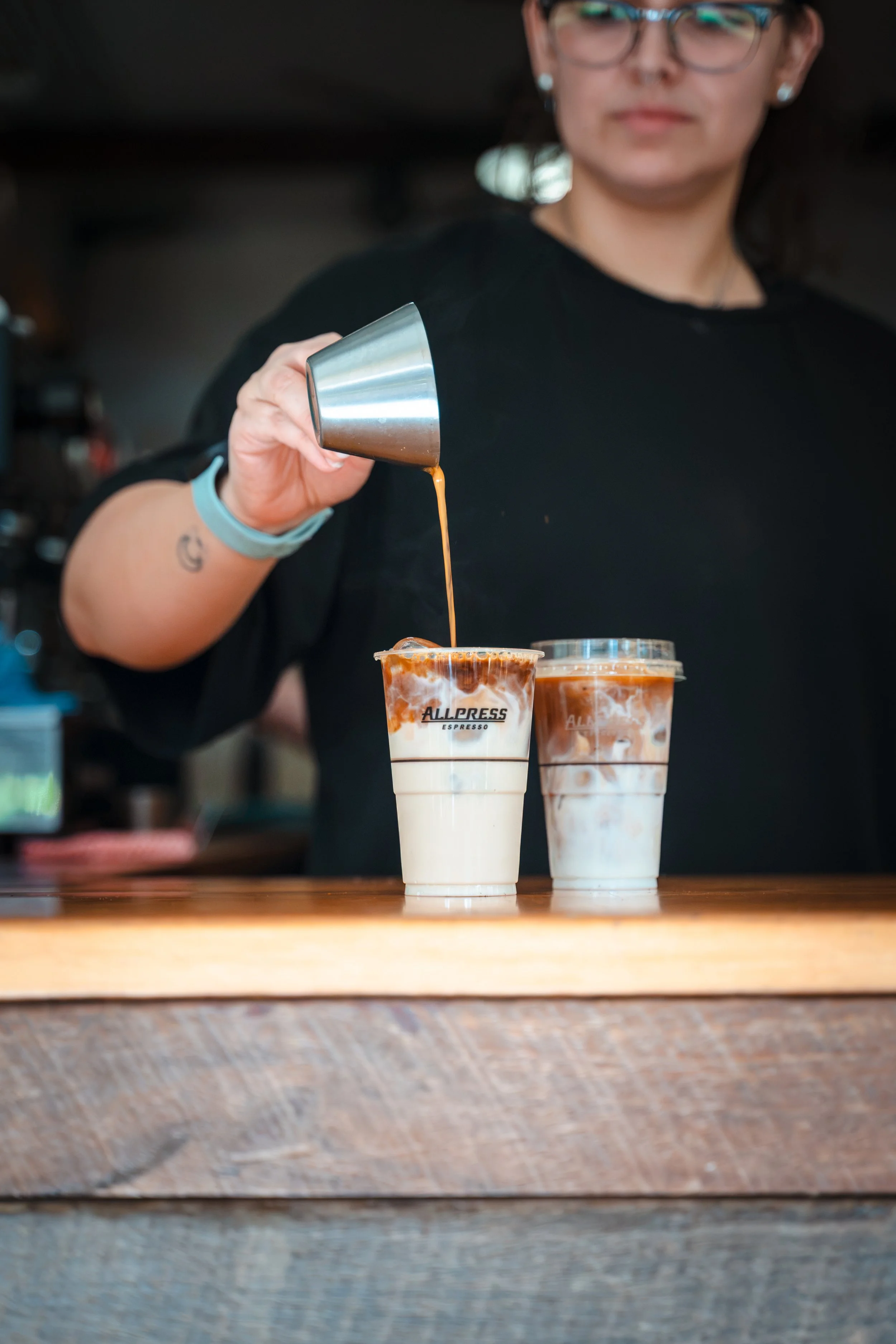 A woman wearing glasses and earrings is pouring caramel syrup into two iced coffee drinks on a wooden counter. The drinks are topped with whipped cream and have a logo that reads 'All Press Espresso'.
