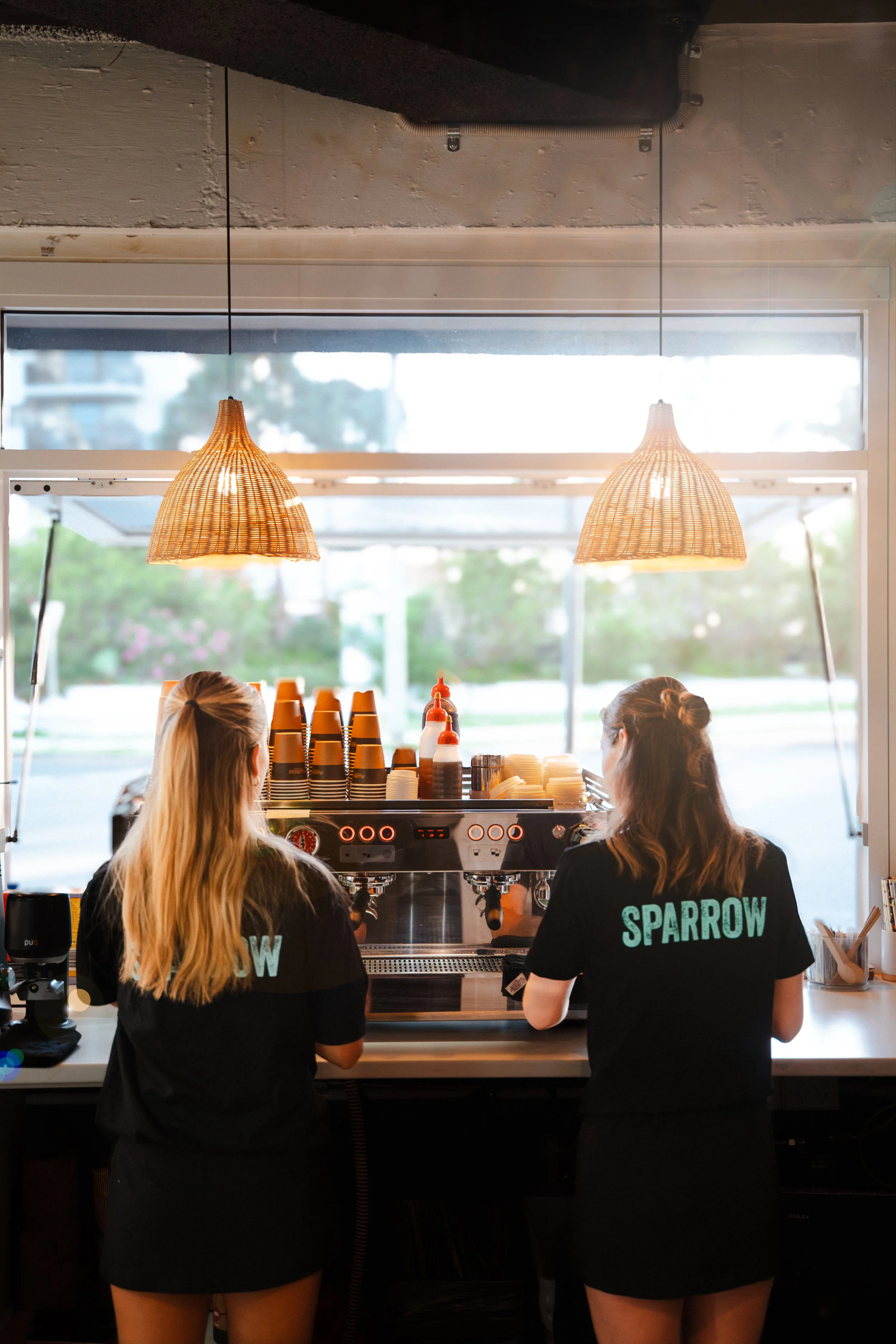 Two women with long hair wearing black shirts with 'SPARROW' written on the back, working behind a coffee bar with espresso machine and cups, seen from behind in a cafe with large window and hanging pendant lights.