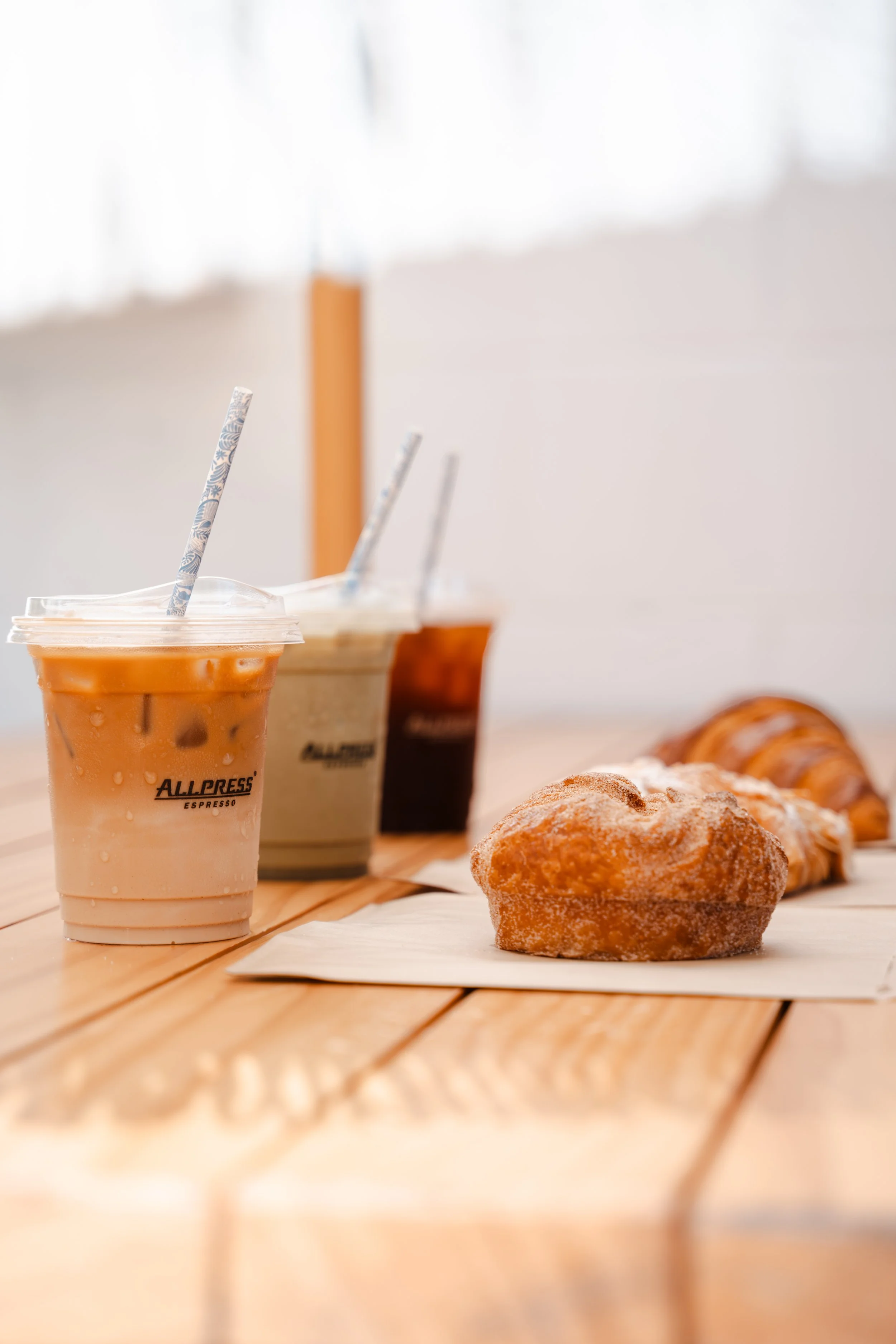 Three cups of iced coffee on a wooden table with a pastry in the foreground, and more pastries in the background.