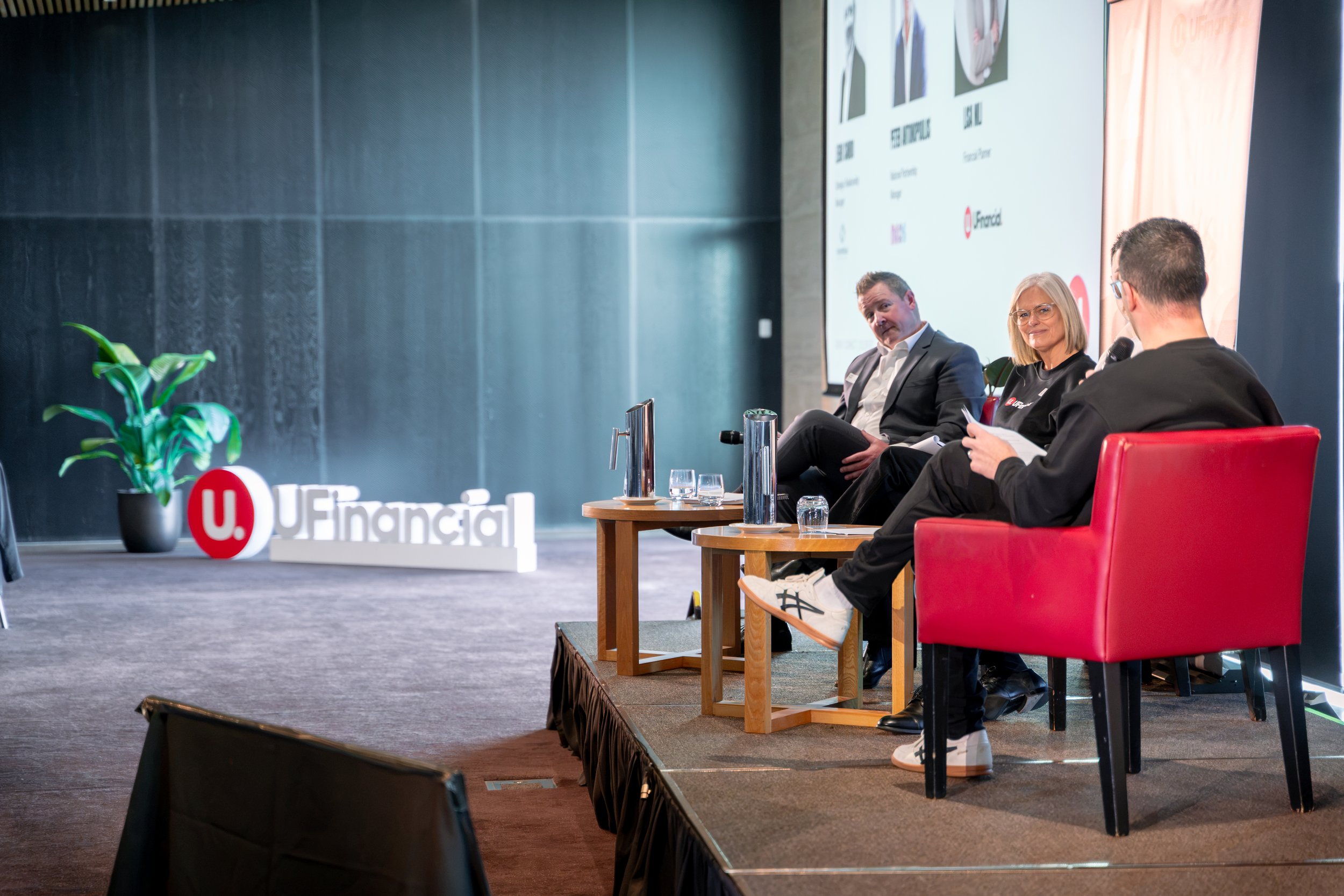 A panel discussion at a conference with three people seated on stage, one man and two women, engaging in conversation. A large screen behind them displays various logos and images. A potted plant and signage for UFinancial are visible in the foreground.