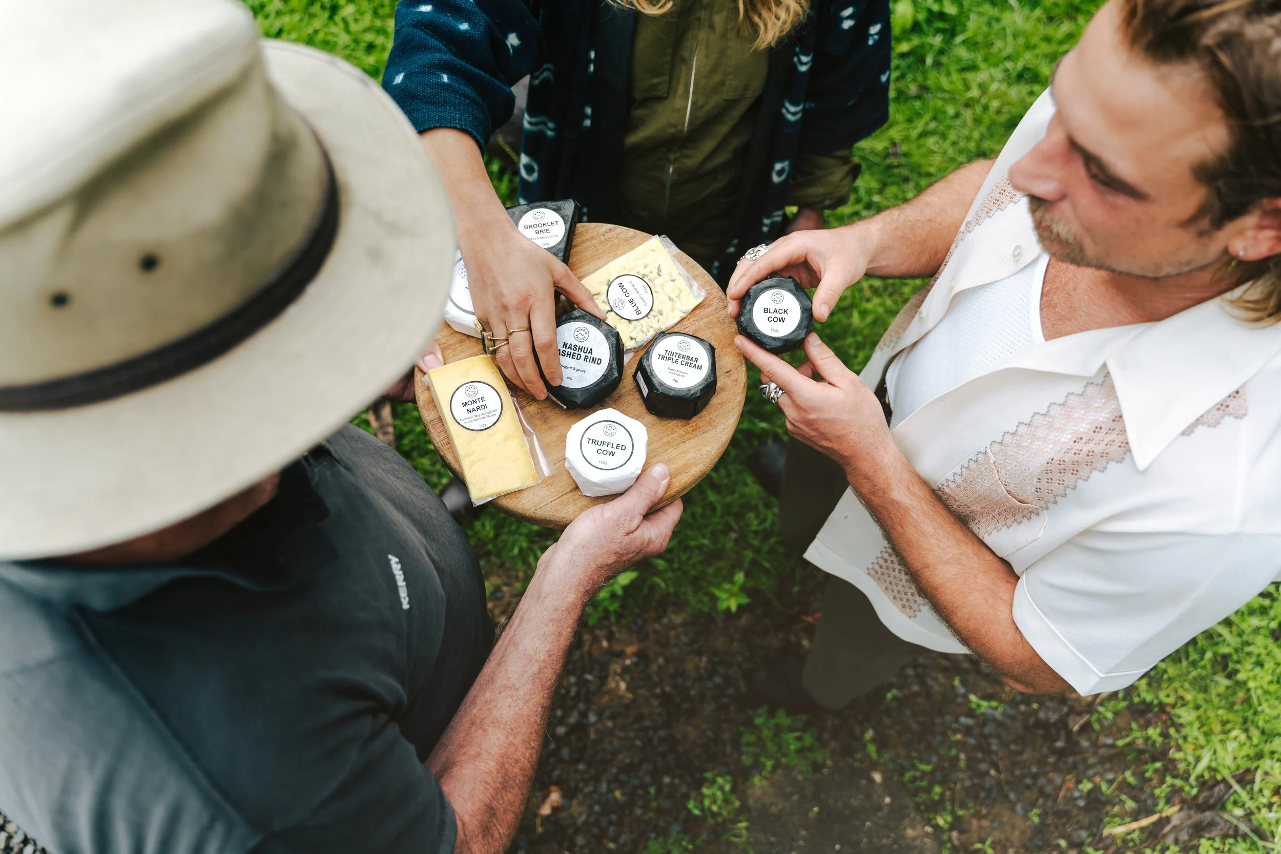 People standing outdoors around a small round wooden table with various cheese products, including Brie, cheddar, and truffle cheese, with one person reaching for a piece of cheese.