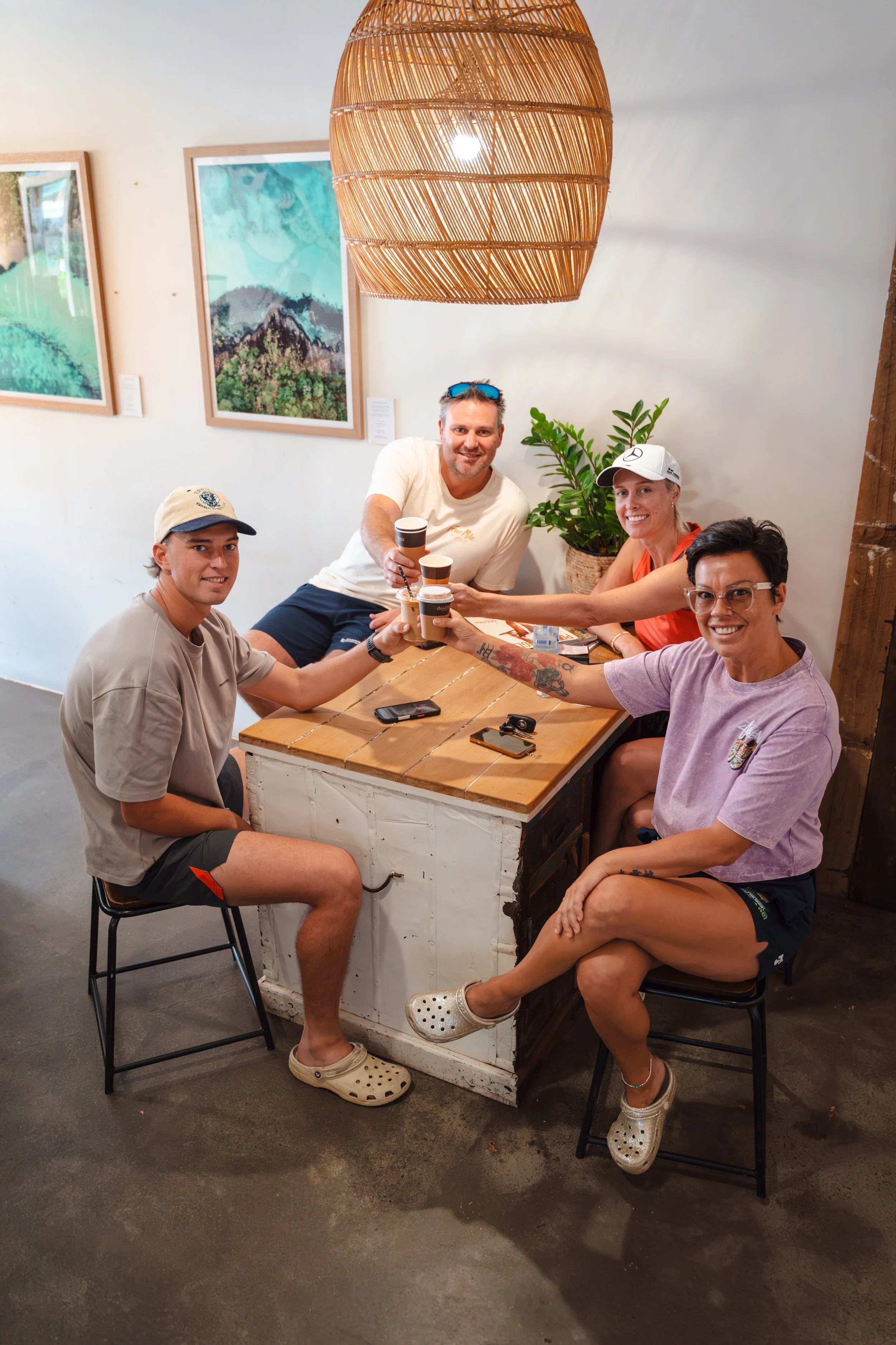 A group of five friends sitting around a rustic wooden table in a cafe, smiling and clinking coffee cups. The cafe has framed artwork on the wall, a large hanging wicker light fixture, and a potted plant.