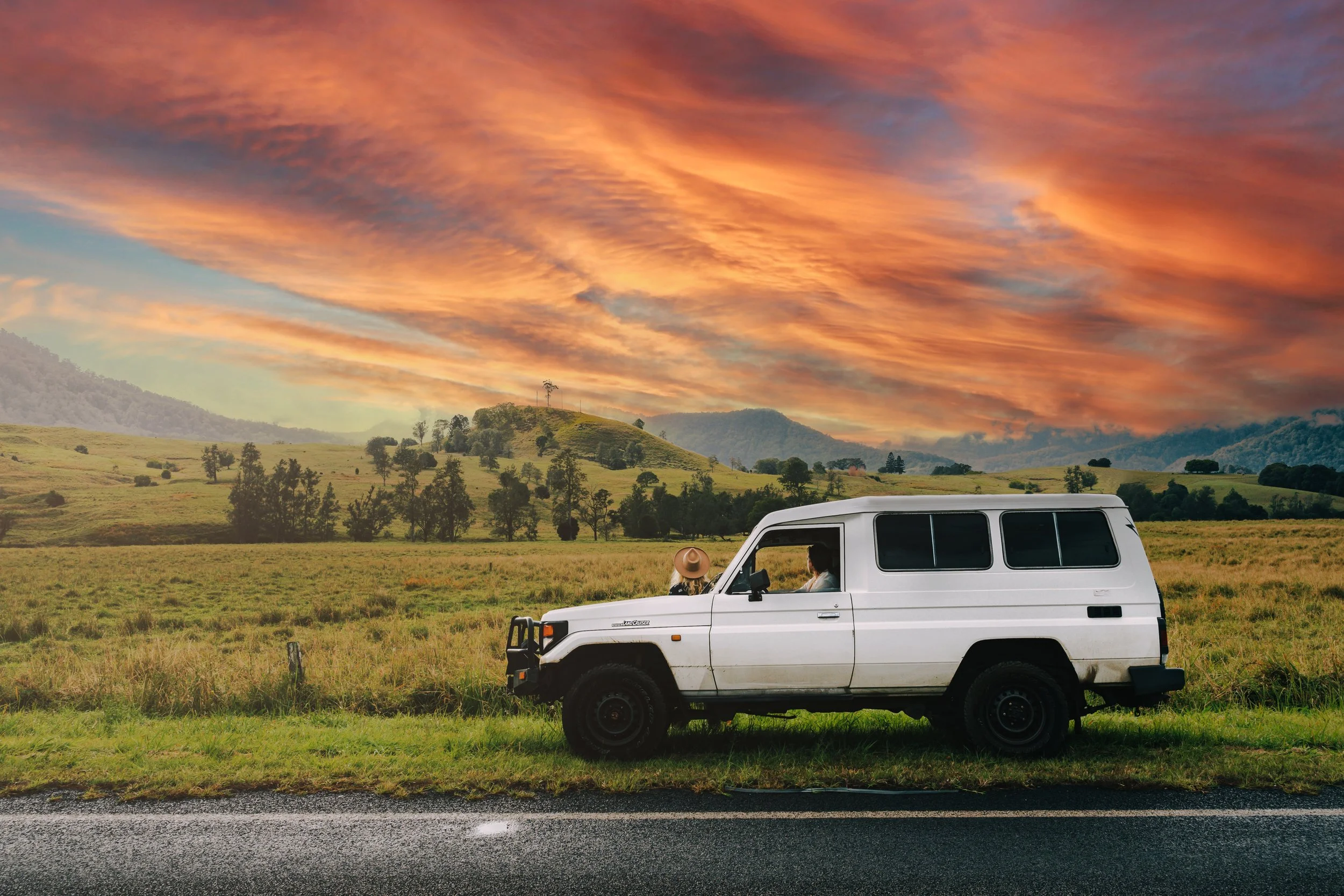 A white off-road vehicle parked on the side of a scenic countryside road. Two people are sitting inside, enjoying the view of the rolling hills and vibrant sunset sky with orange and purple clouds.