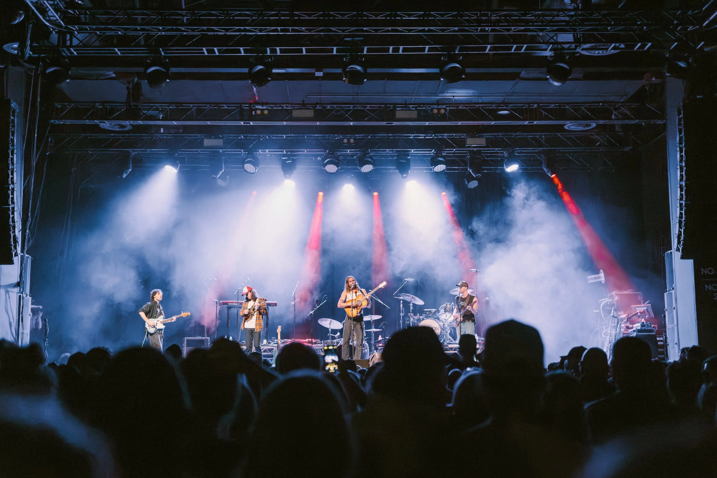 A live band performing on stage with multiple musicians playing guitars and drums, bright stage lights shining down, and an audience watching in the foreground.