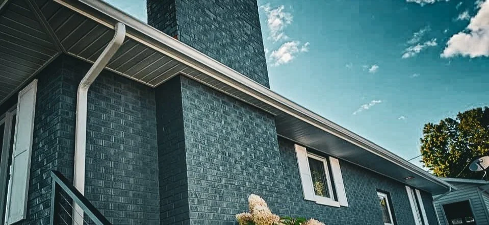 Close-up of a modern house with a brick exterior, white window frames, and a white gutter system under a partly cloudy sky.