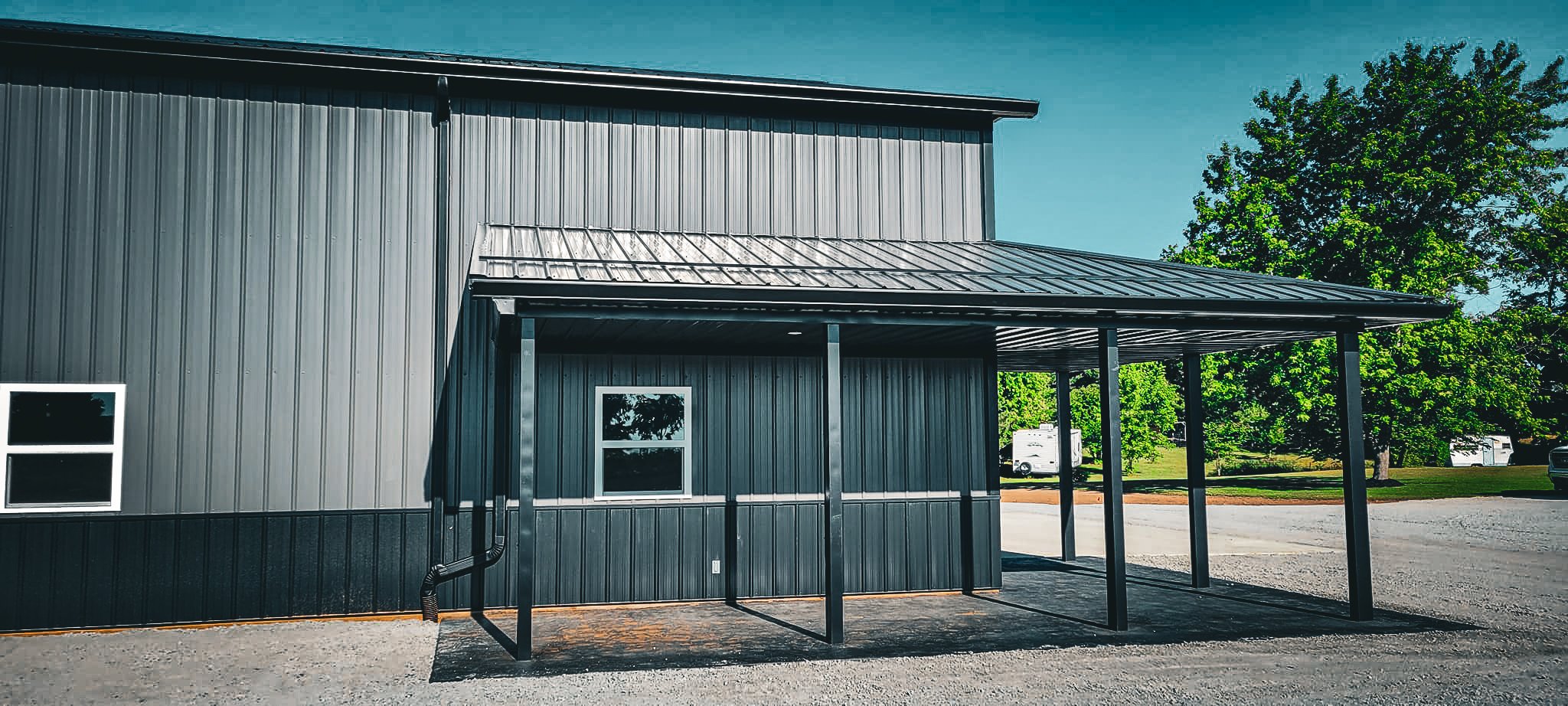 Steel building with a metal awning, two small windows, black vertical siding, and surrounded by trees and parked trailers.