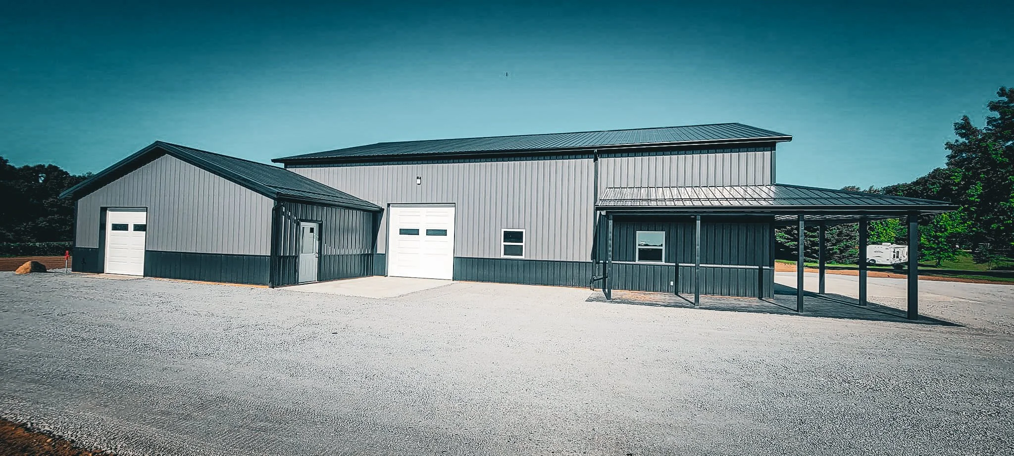 A large gray metal building with multiple sections, including small garage doors and a covered area, on a gravel lot with trees in the background.
