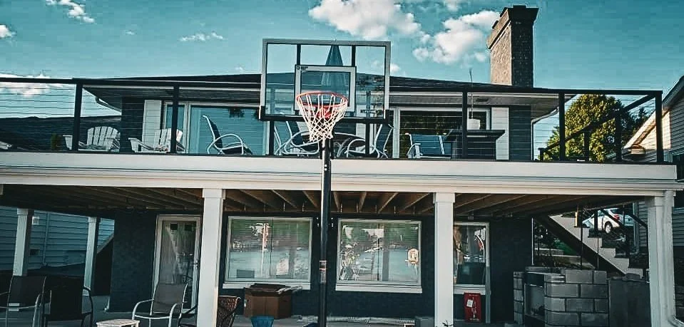Backyard deck with a basketball hoop mounted on the railing, chairs, and patio furniture, with a house and blue sky in the background.