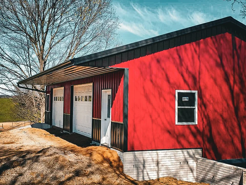Red barn with black roof, two garage doors, small window, set against a sunny sky with leafless trees.