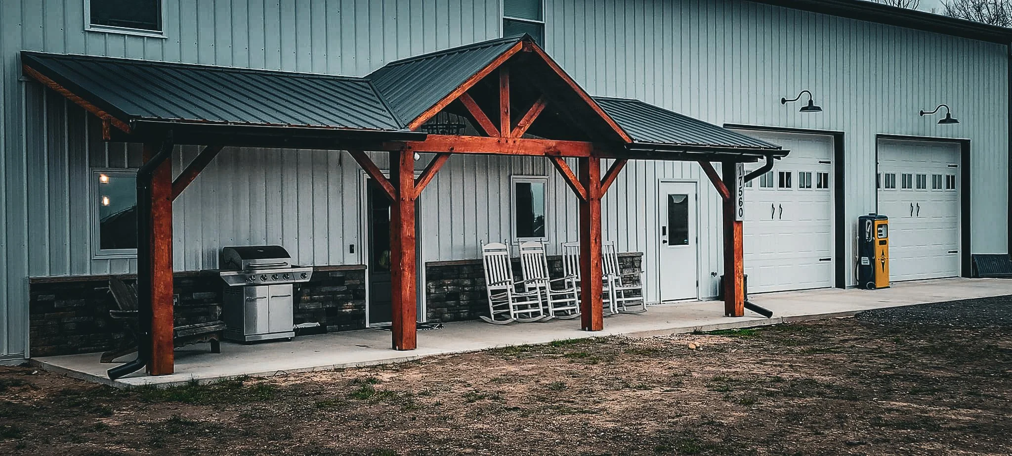 Metal building with a decorated entrance, white garage doors, outdoor chairs, gas pump, and grill, with gravel ground in front.