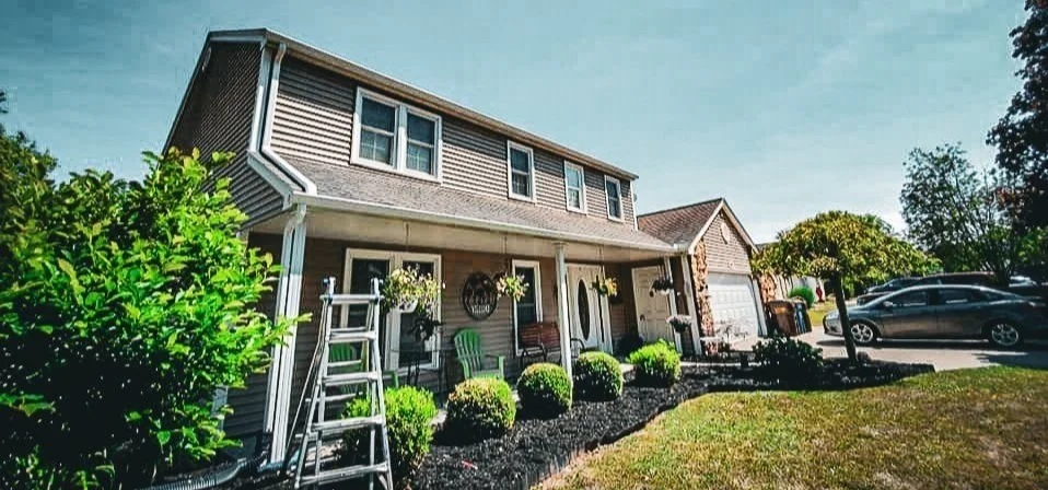 A two-story house with a front porch decorated with plants and hanging baskets, a driveway with cars, and a well-maintained front yard with bushes and trees.