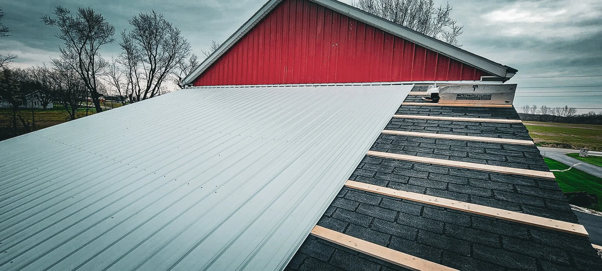 A partially replaced roof on a barn, with black shingles and a new white metal roofing section being installed.