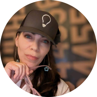 a brown-haired woman with blue eyes, a dark grey trucker hat with a light bulb graphic, holding pink glasses in front of an abstract marquis letter backdrop