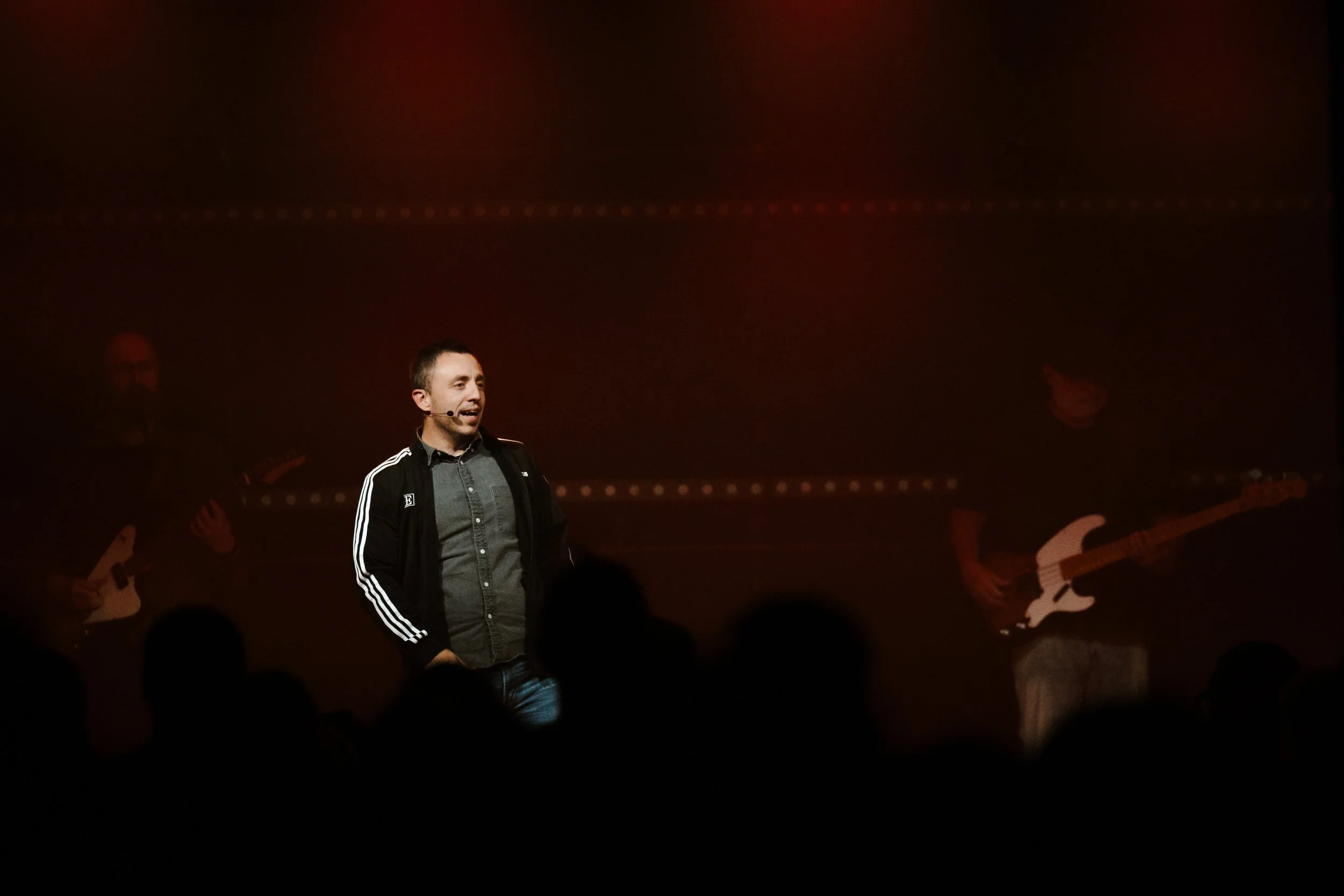 Man speaking or singing on stage with two musicians playing guitars in the background and an audience in the foreground.