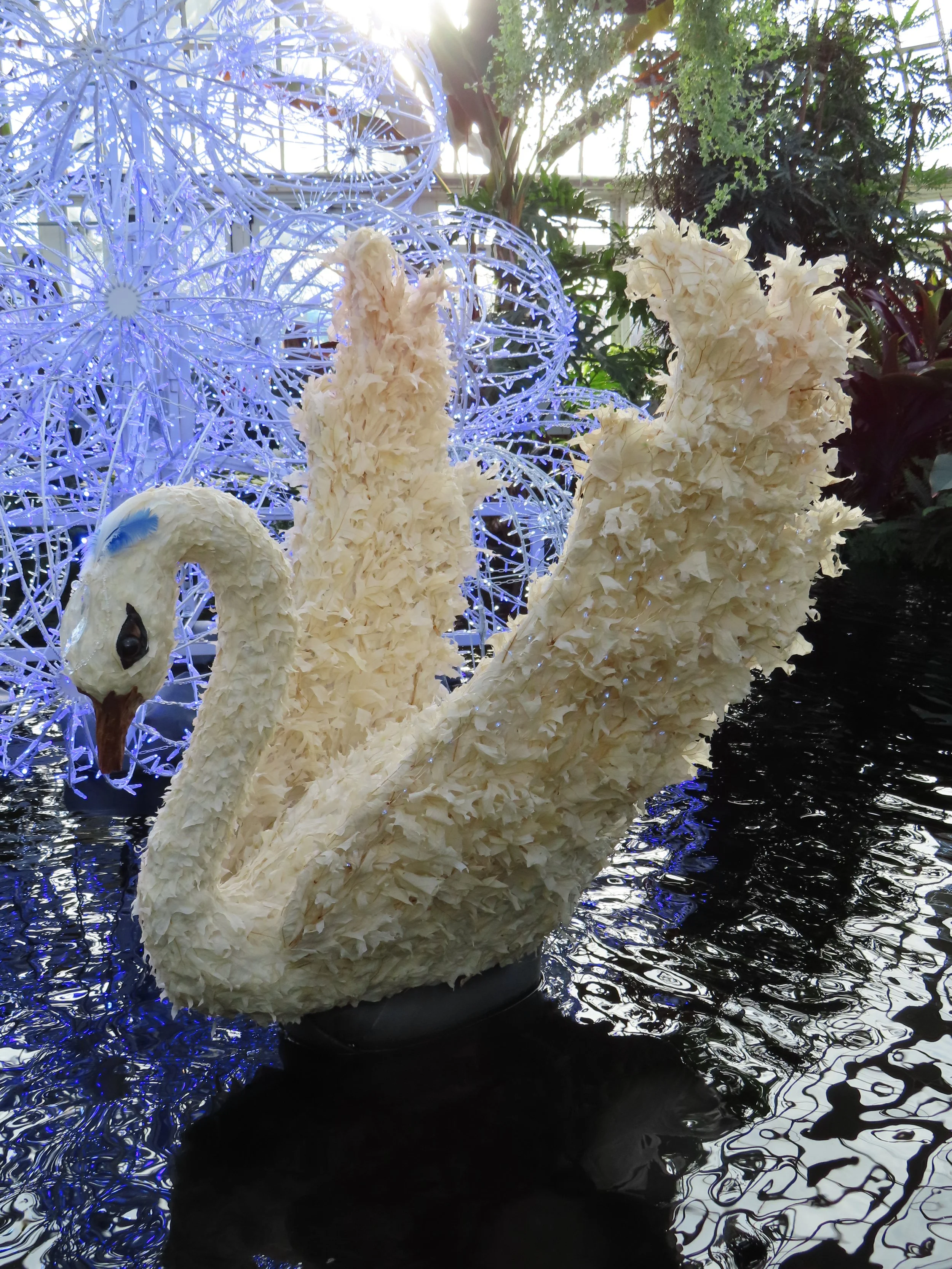 Decorative white swan sculpture on water with intricate blue illuminated spherical structures and green plants in the background.