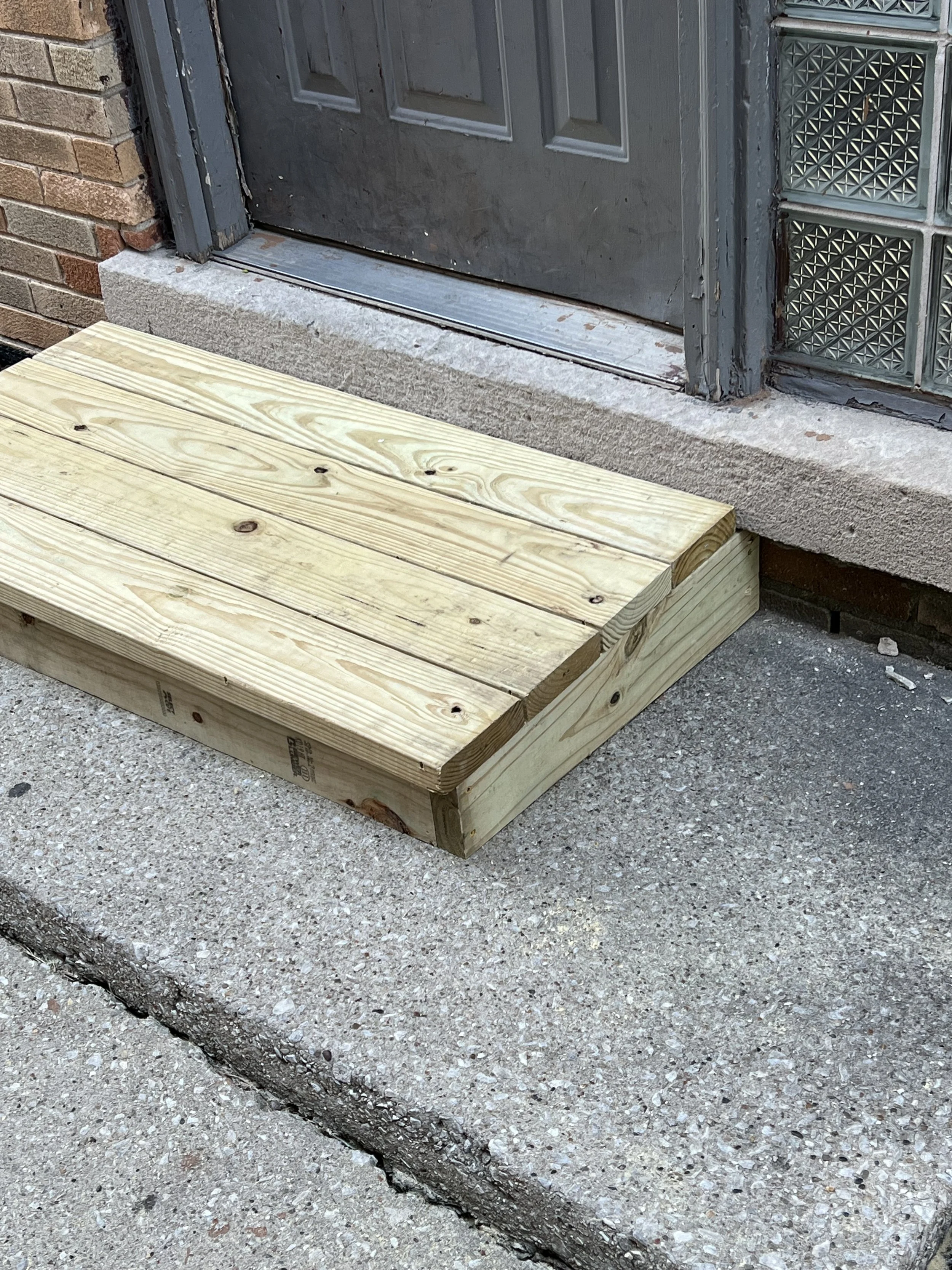 A newly built wooden step in front of a gray door with glass bricks on the side, set on a concrete sidewalk with a brick wall to the left.