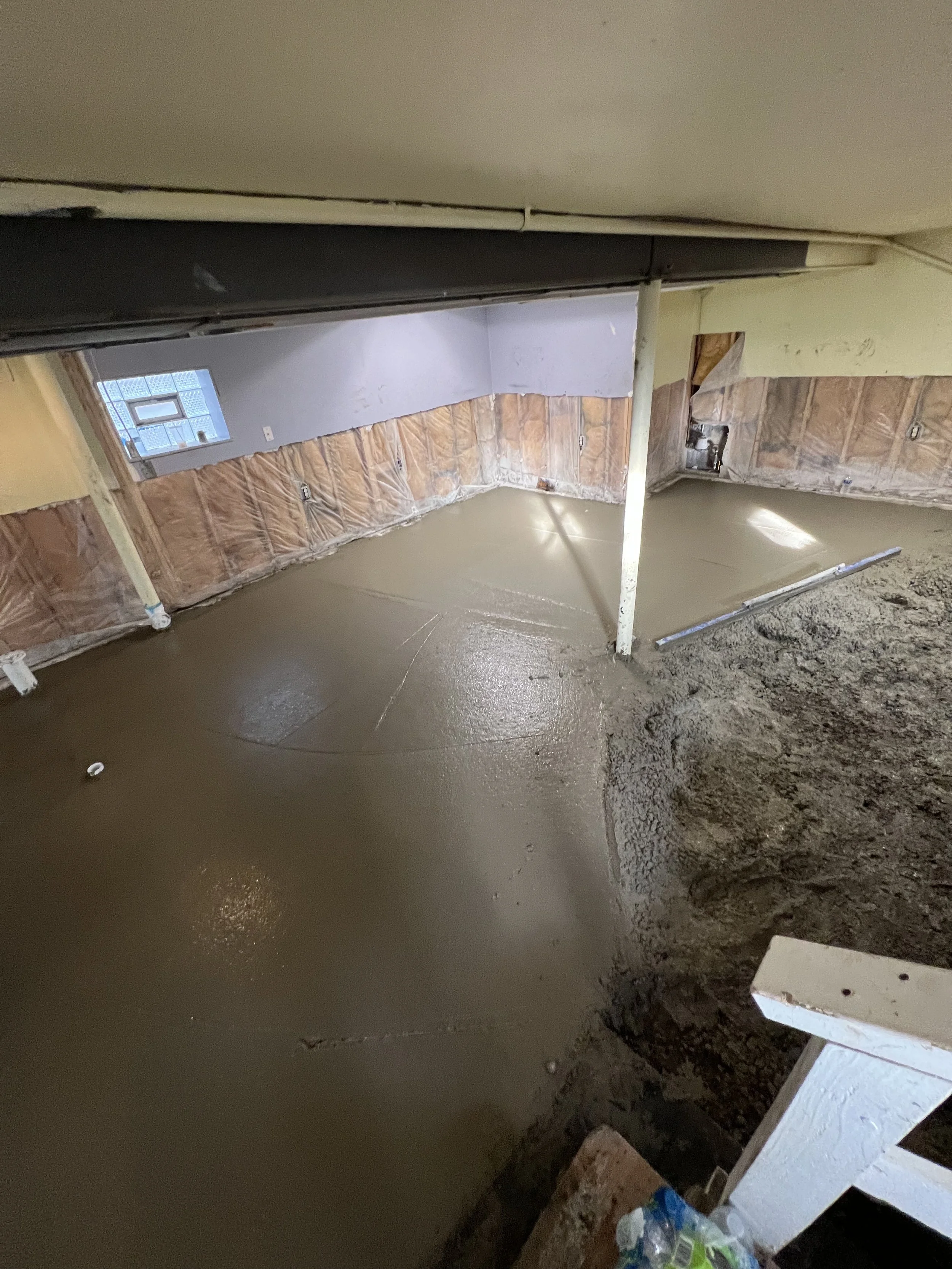 A basement under construction with freshly poured concrete flooring, exposed insulation, a small window, and construction materials on the floor.