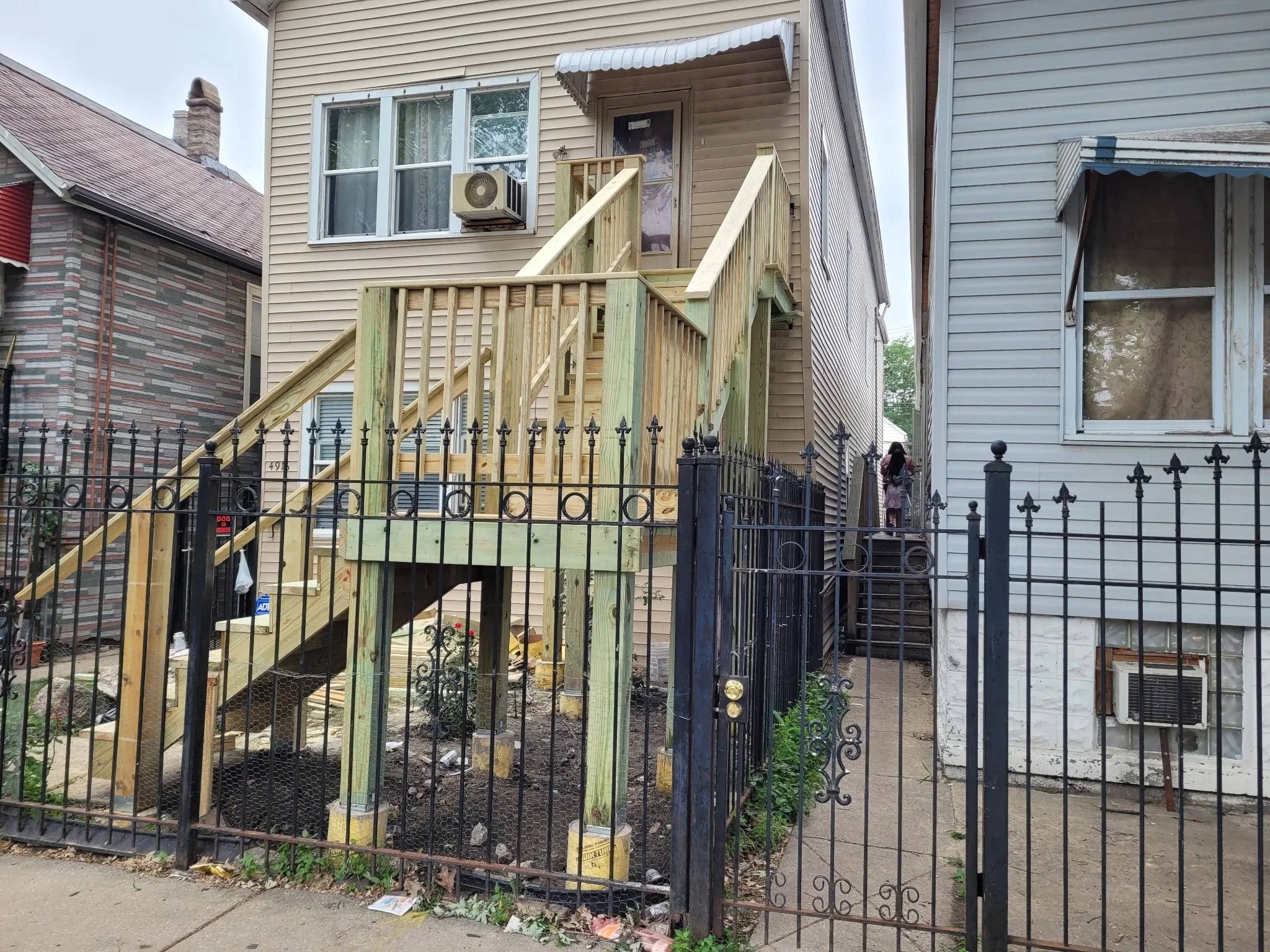 Two houses with a fenced yard, one house has a new wooden staircase leading to a door. A person is walking up the stairs.