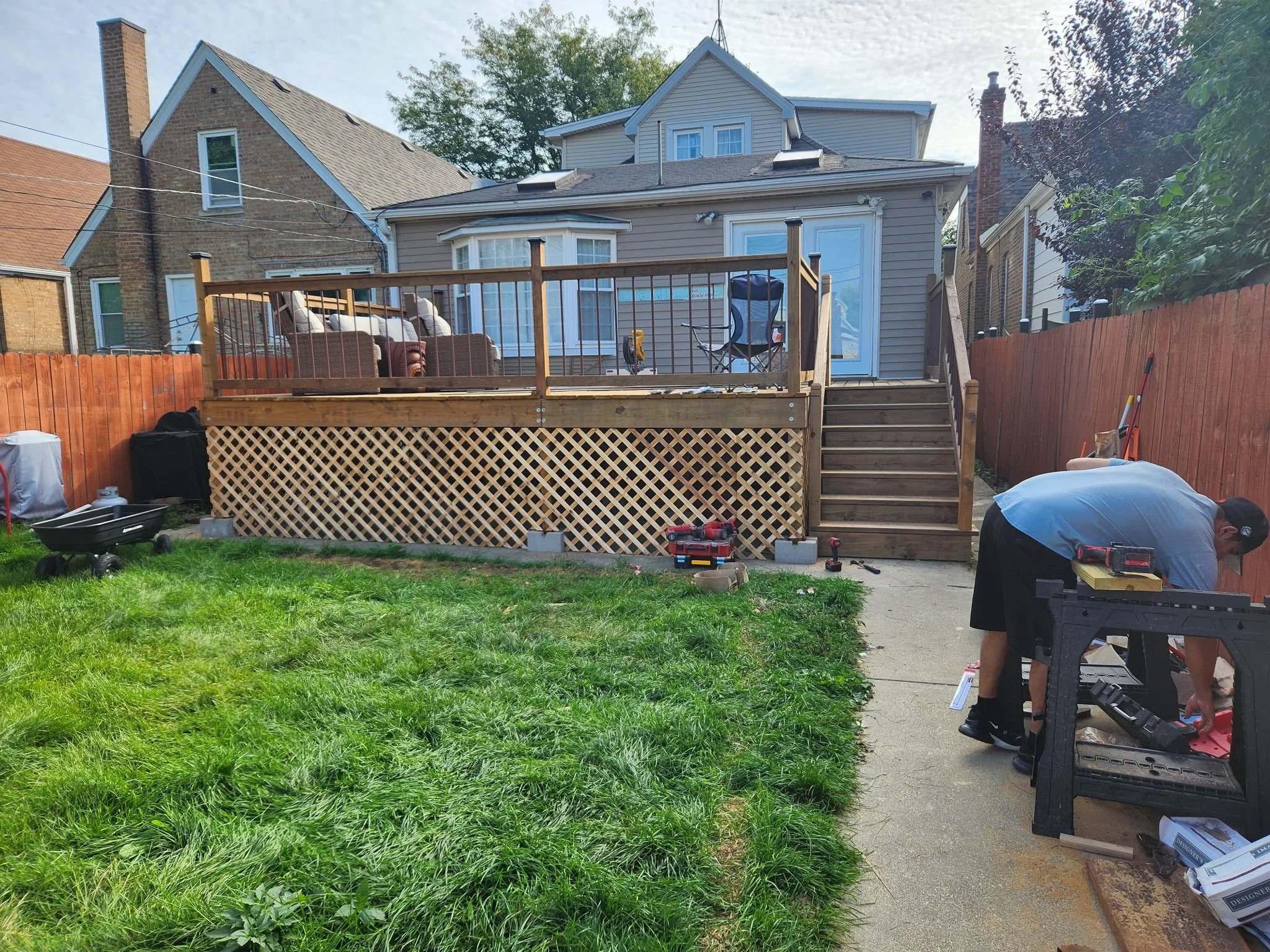 A man working on a backyard deck construction with tools, near a grassy yard, wooden fencing, and neighboring houses in the background.