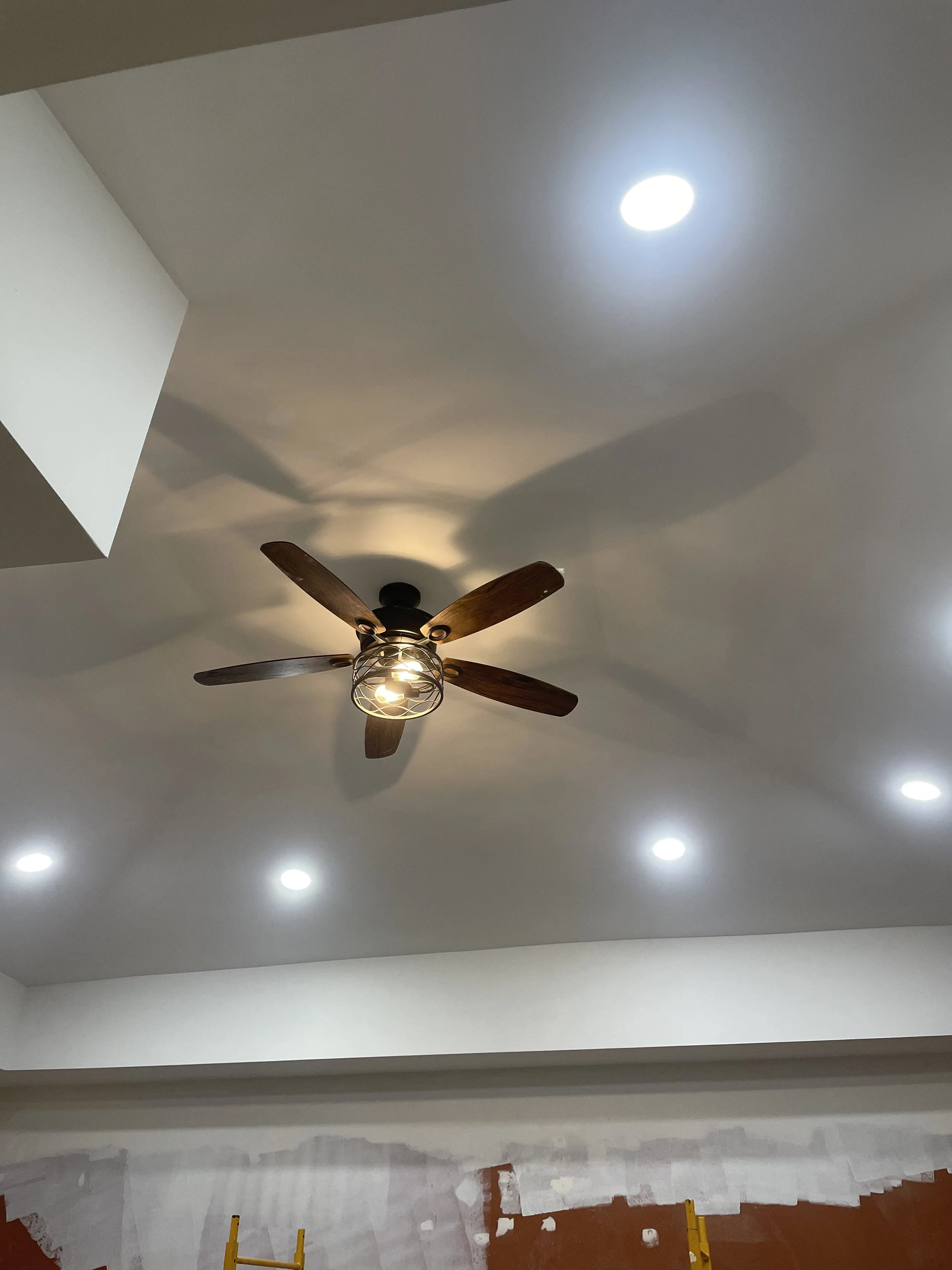 Ceiling with a ceiling fan made of wood and metal, illuminated by multiple recessed lights, with some construction work visible below.