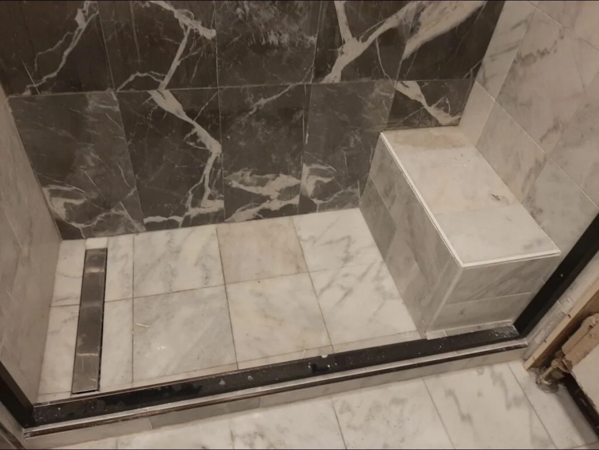 Inside a shower with marble tiles, a built-in marble bench, and a metal drain at the bottom.