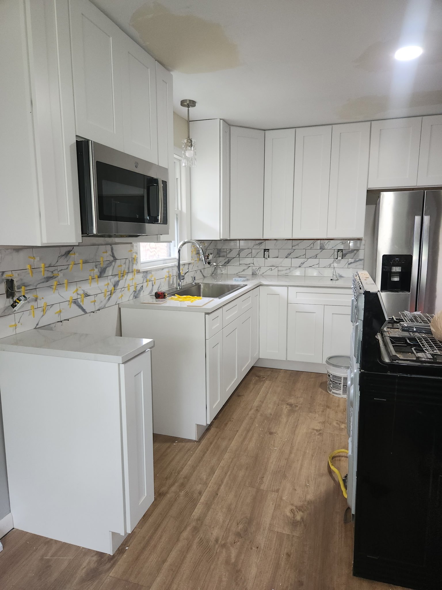 Kitchen with white cabinets, marble backsplash, stainless steel appliances, wooden floor, yellow clothespins and tools on countertops, old and new cabinets, under renovation.