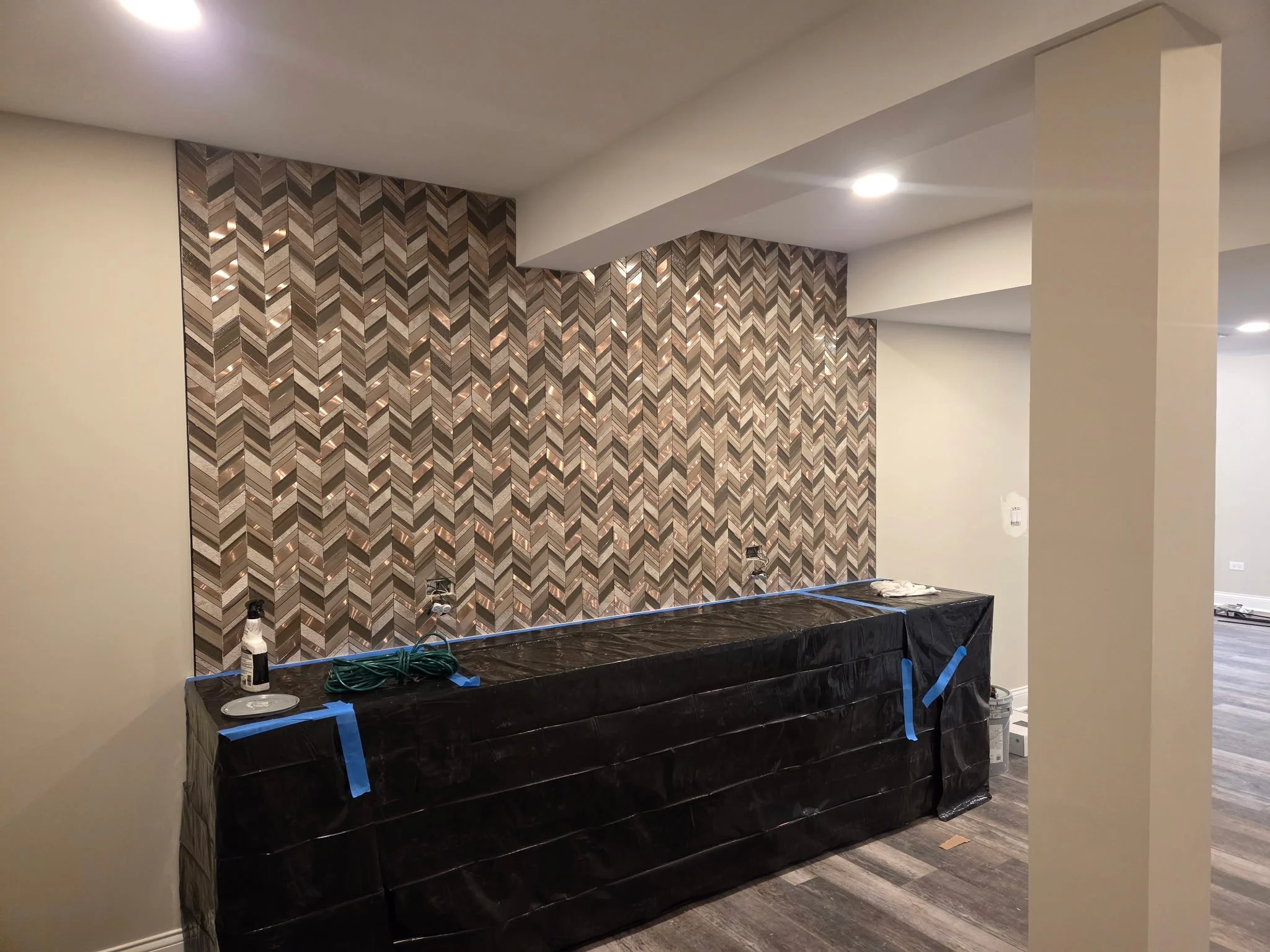 Under construction or renovation area with a dark countertop or cabinet covered with black plastic, tape, and cleaning supplies, against a patterned wall with geometric tile in a room with recessed lighting and wood-look flooring.