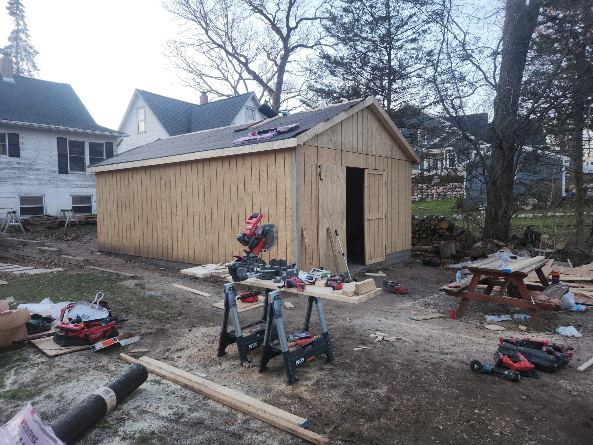 A partially constructed wooden shed in a backyard, with tools and materials scattered around, and houses in the background.