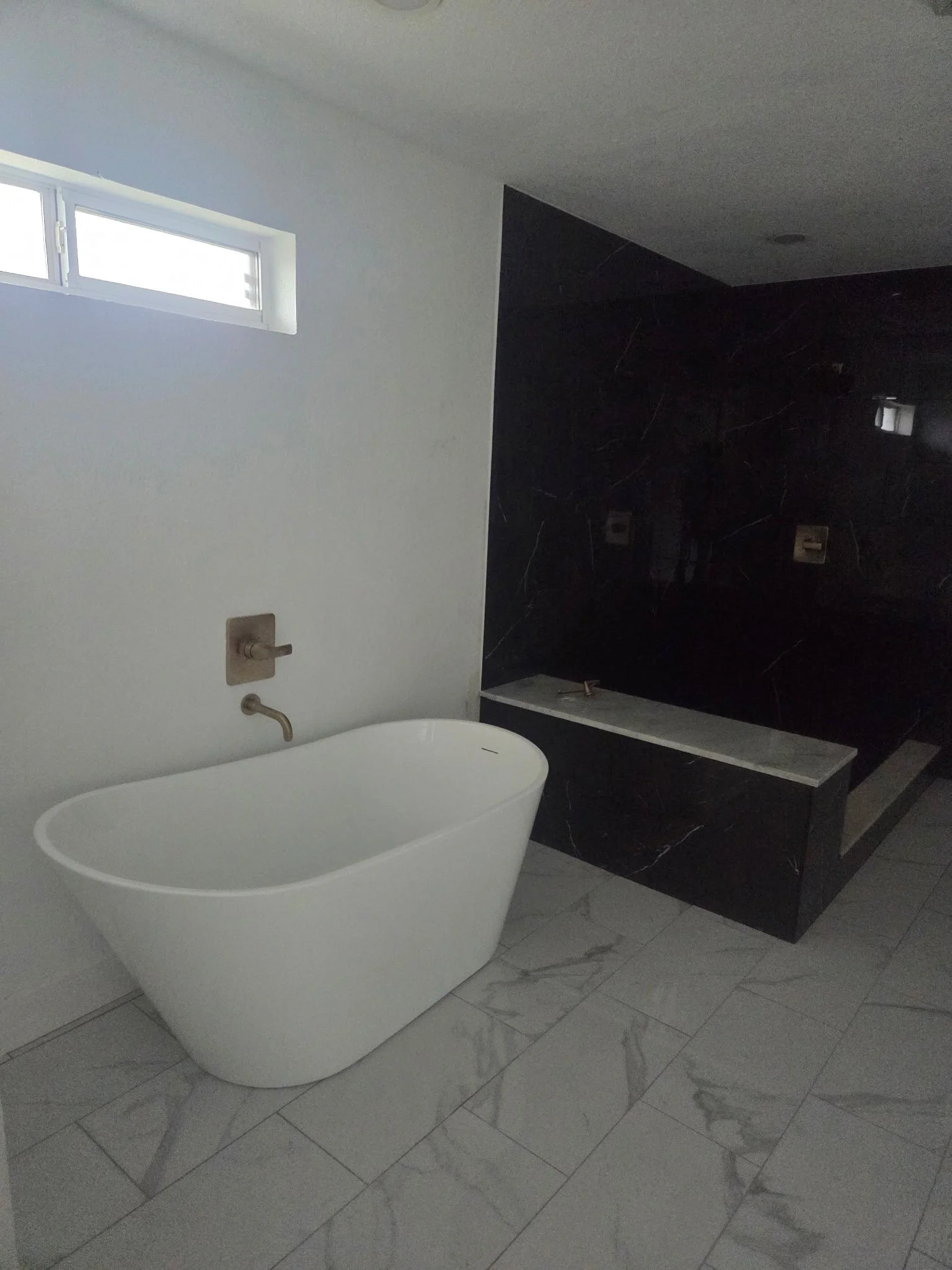 Modern bathroom with a white freestanding bathtub, gray marble floor tiles, a window, and black marble accent wall.