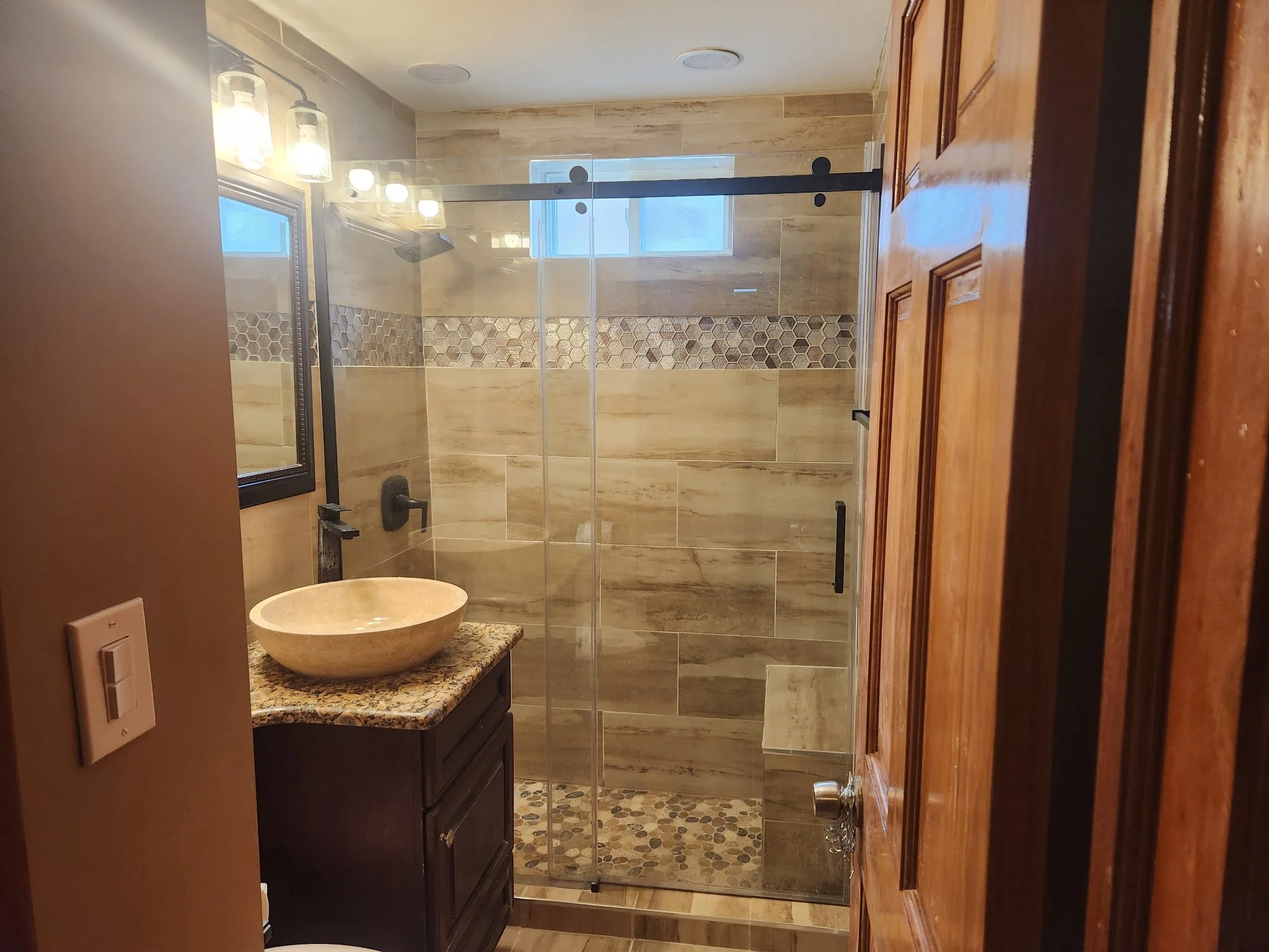 Bathroom with wood door, granite countertop with vessel sink, mirror, and lighting fixtures, walk-in shower with glass door, tan tiles, a decorative strip of hexagon tiles, and a small window.