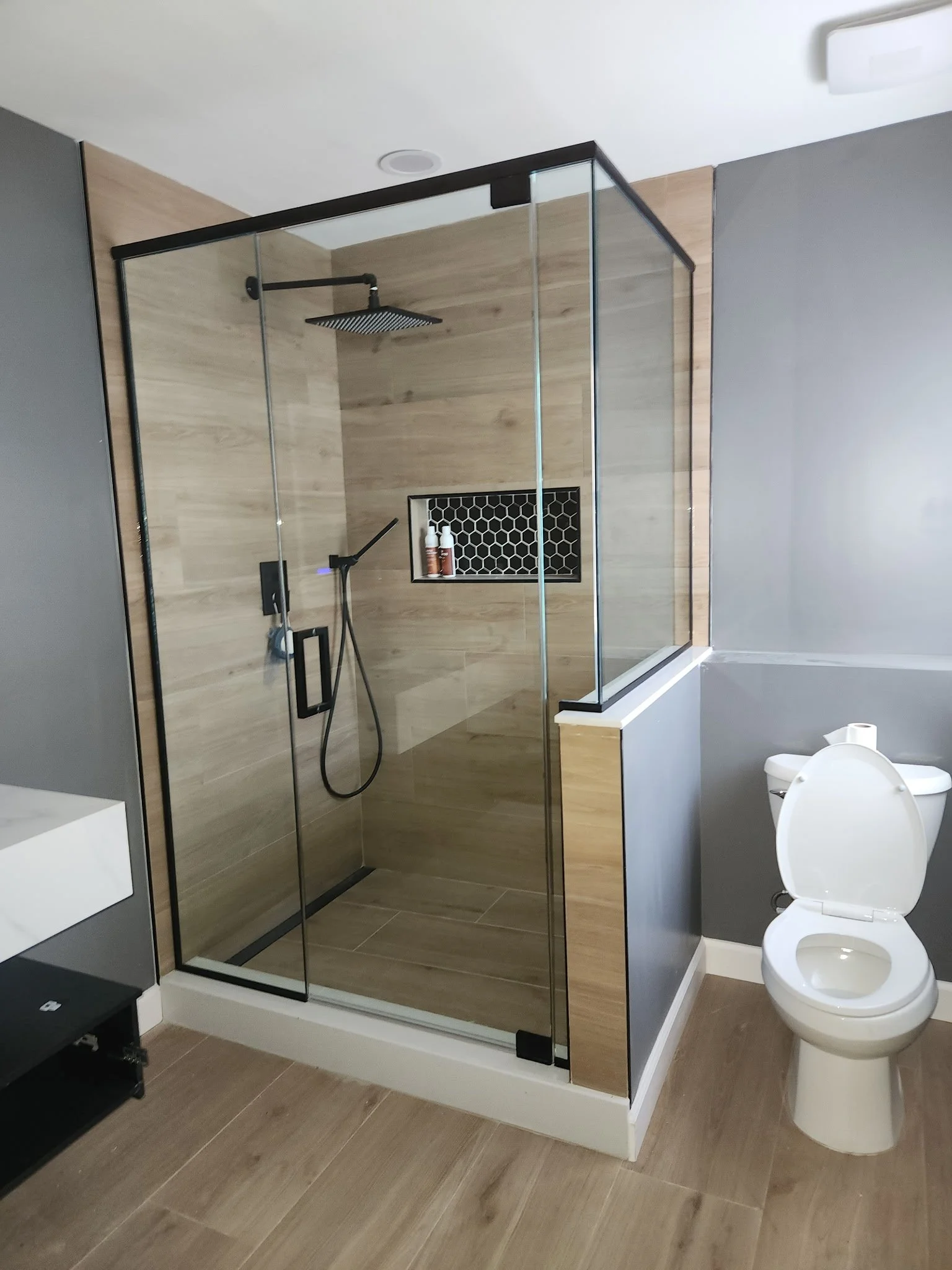 Modern bathroom with walk-in glass shower, black shower fixtures, built-in niche with bottles, gray walls, wooden floor, and a white toilet.