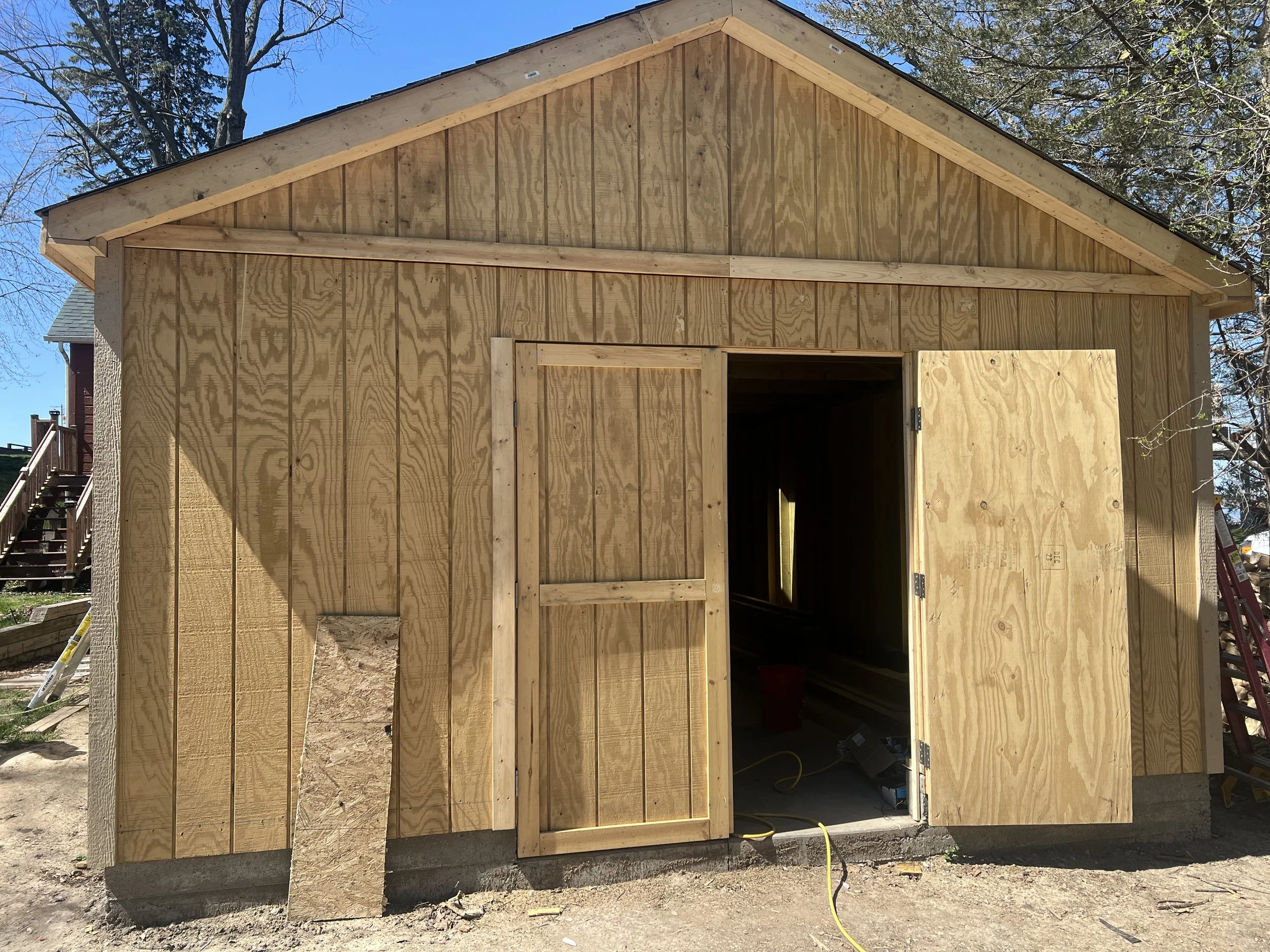 Frame of a small wooden shed being built with plywood walls and a roof, with an open door and a piece of plywood leaning against the wall.