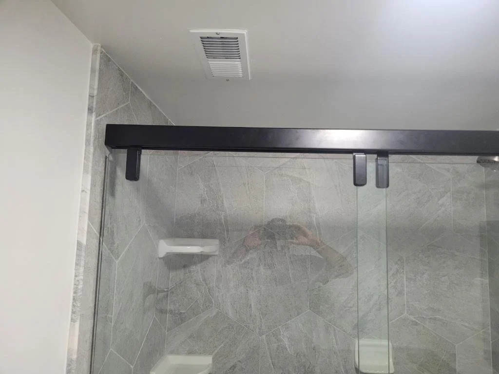 Glass shower door with black metal track, gray tiled shower walls, shower soap dish, vent on the ceiling, and a reflection of a person taking the photo.