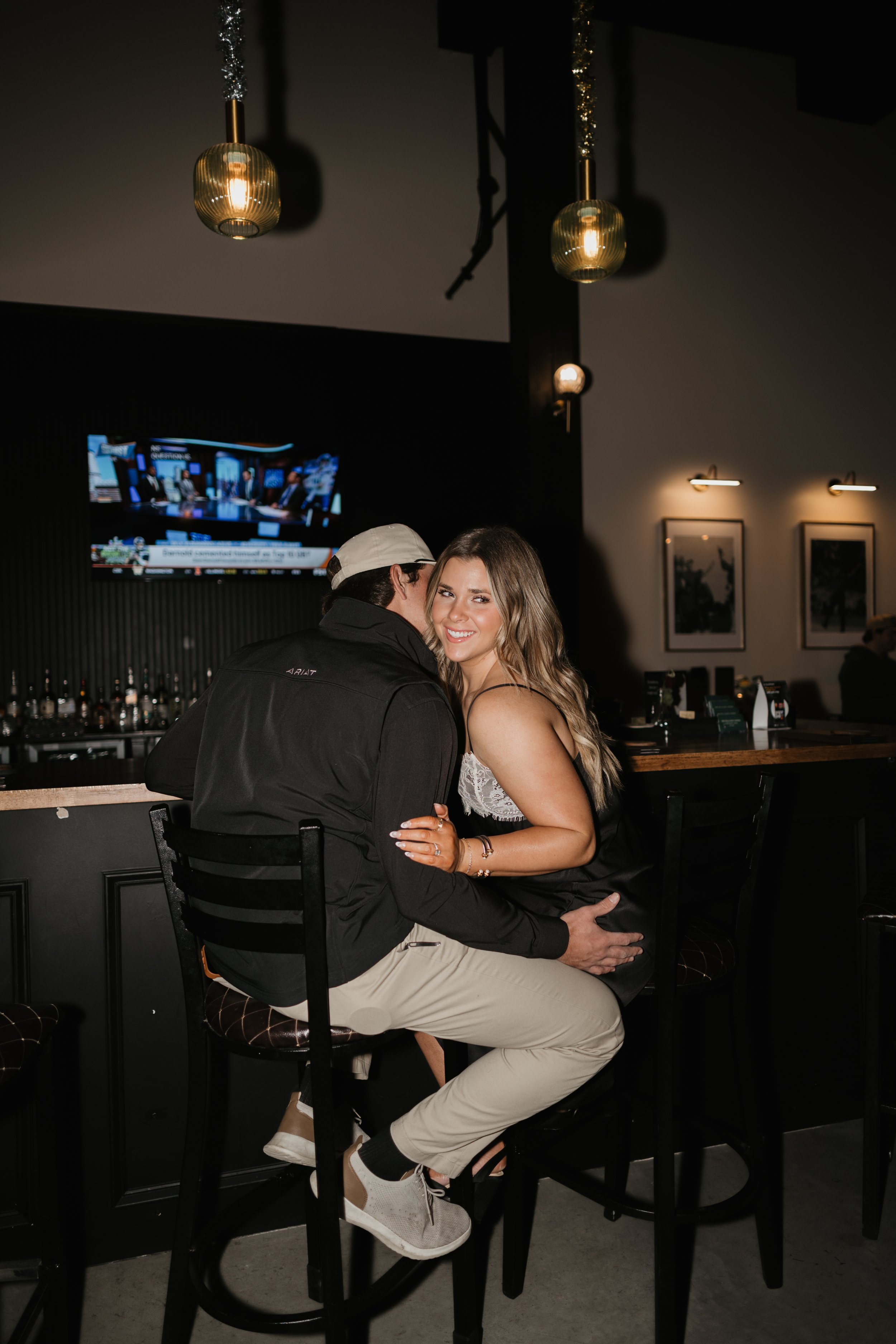 A smiling woman and a man sitting on barstools at a bar, with the woman smiling at the camera. The woman has long blonde hair and is wearing a black dress with white lace detail. The man is wearing a black jacket and a white cap. There are pendant lights hanging from the ceiling, artwork on the wall, and a television screen in the background.