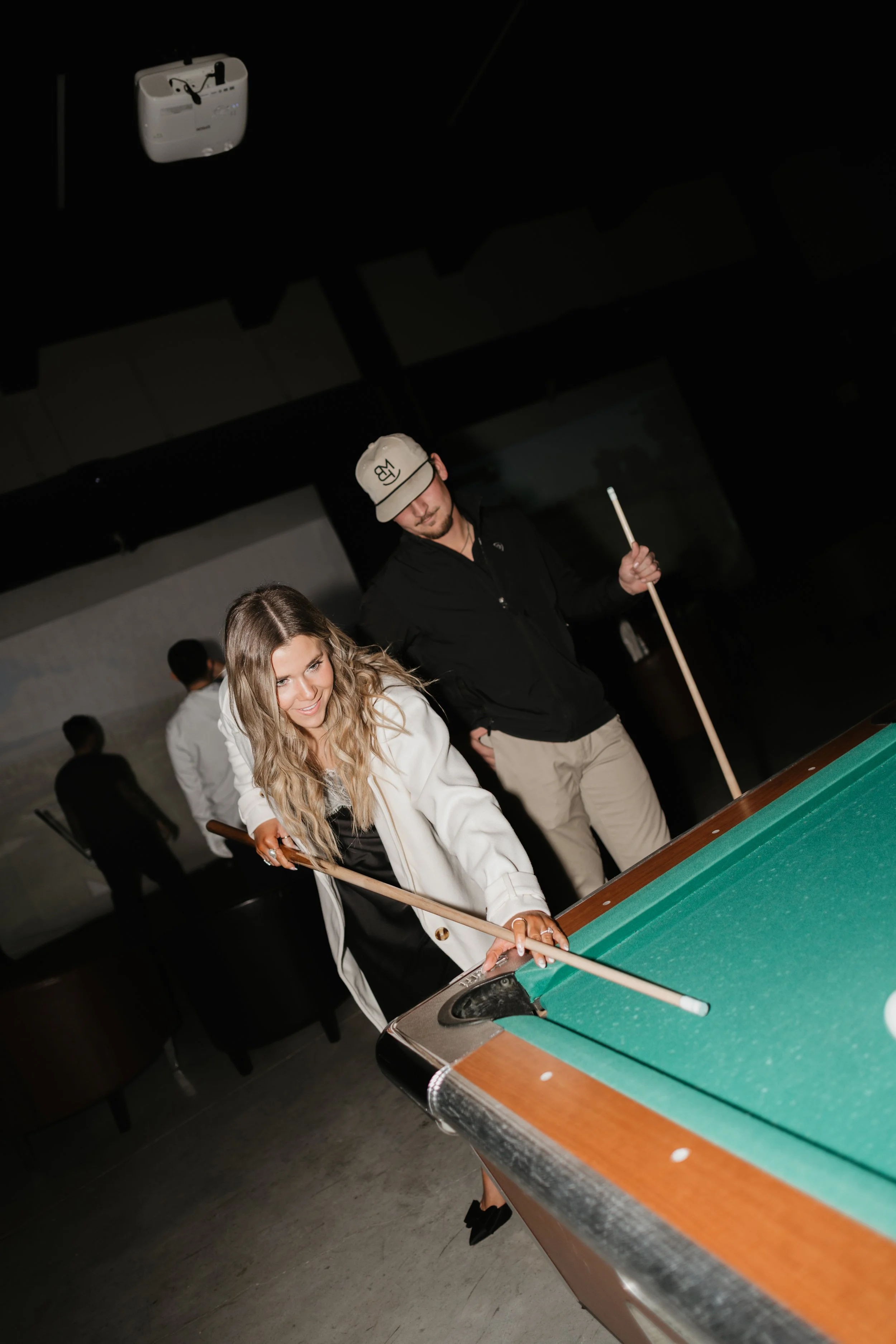 Woman and man playing pool in a dimly lit room with other people in the background.