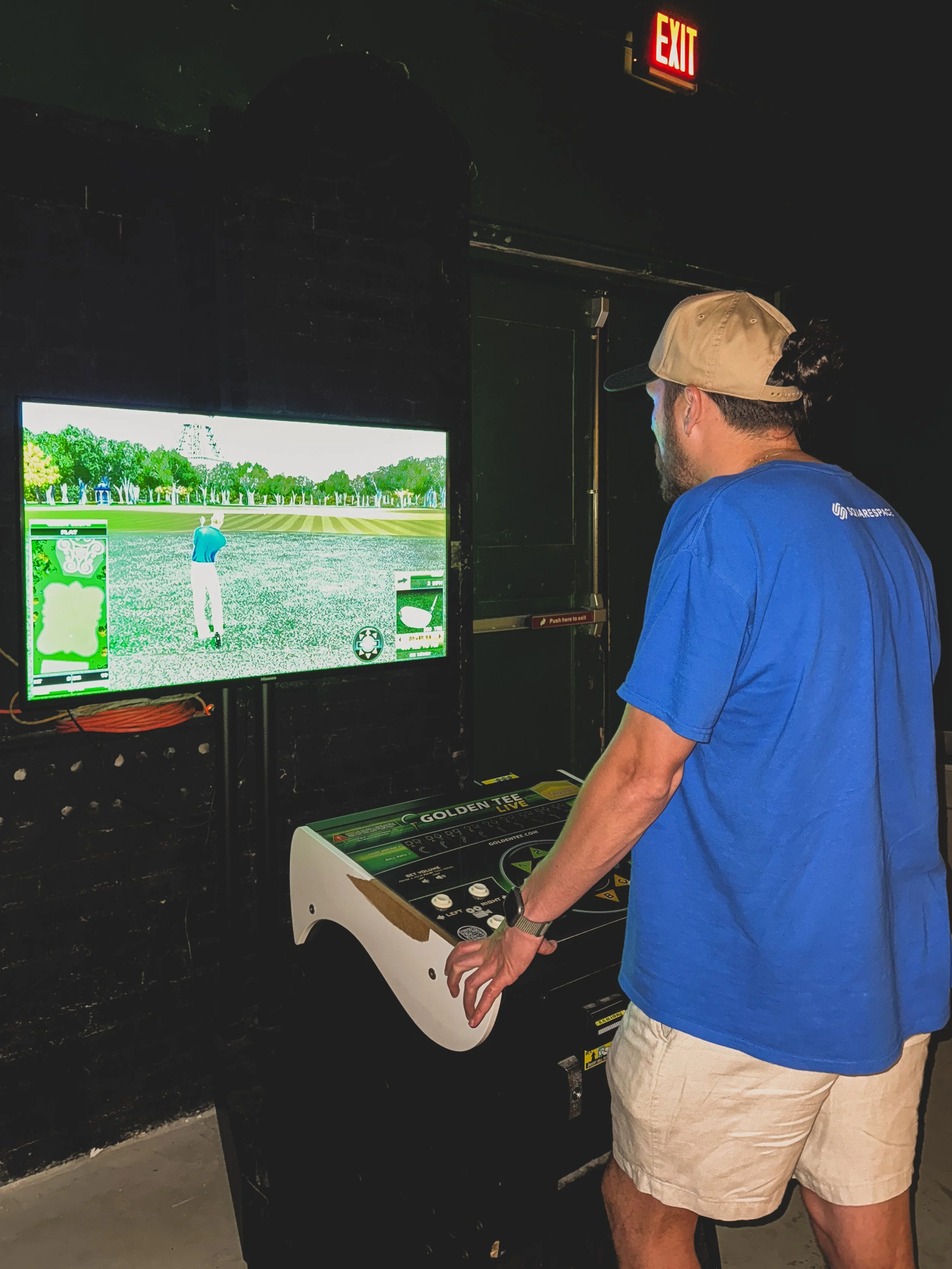A man playing the golf video game, Golden Tee on an arcade machine in a dark room. He is wearing a blue shirt, beige shorts, and a tan cap, and standing in front of a large screen displaying a golf course.