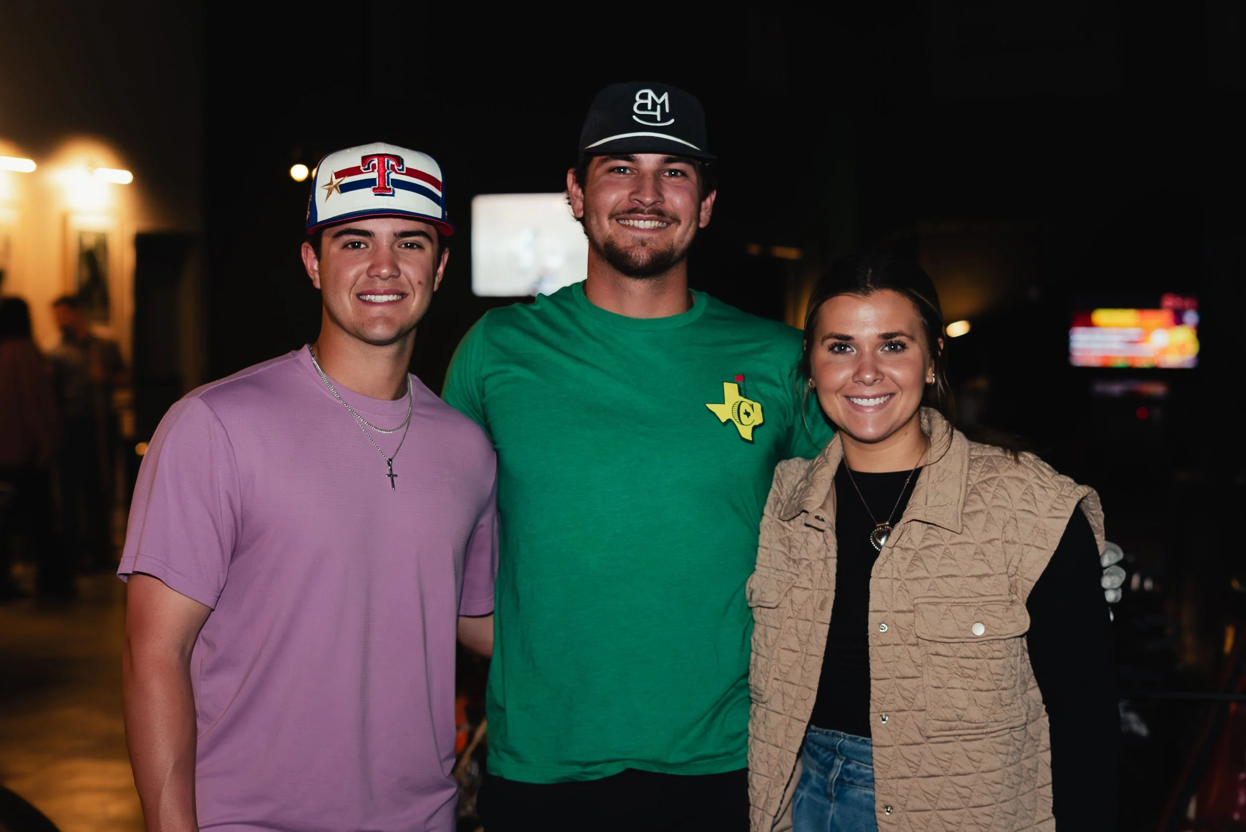 Three young adults standing together indoors at night, smiling at the camera. Two men and one woman, dressed casually, with a blurred background including lights and electronic screens.
