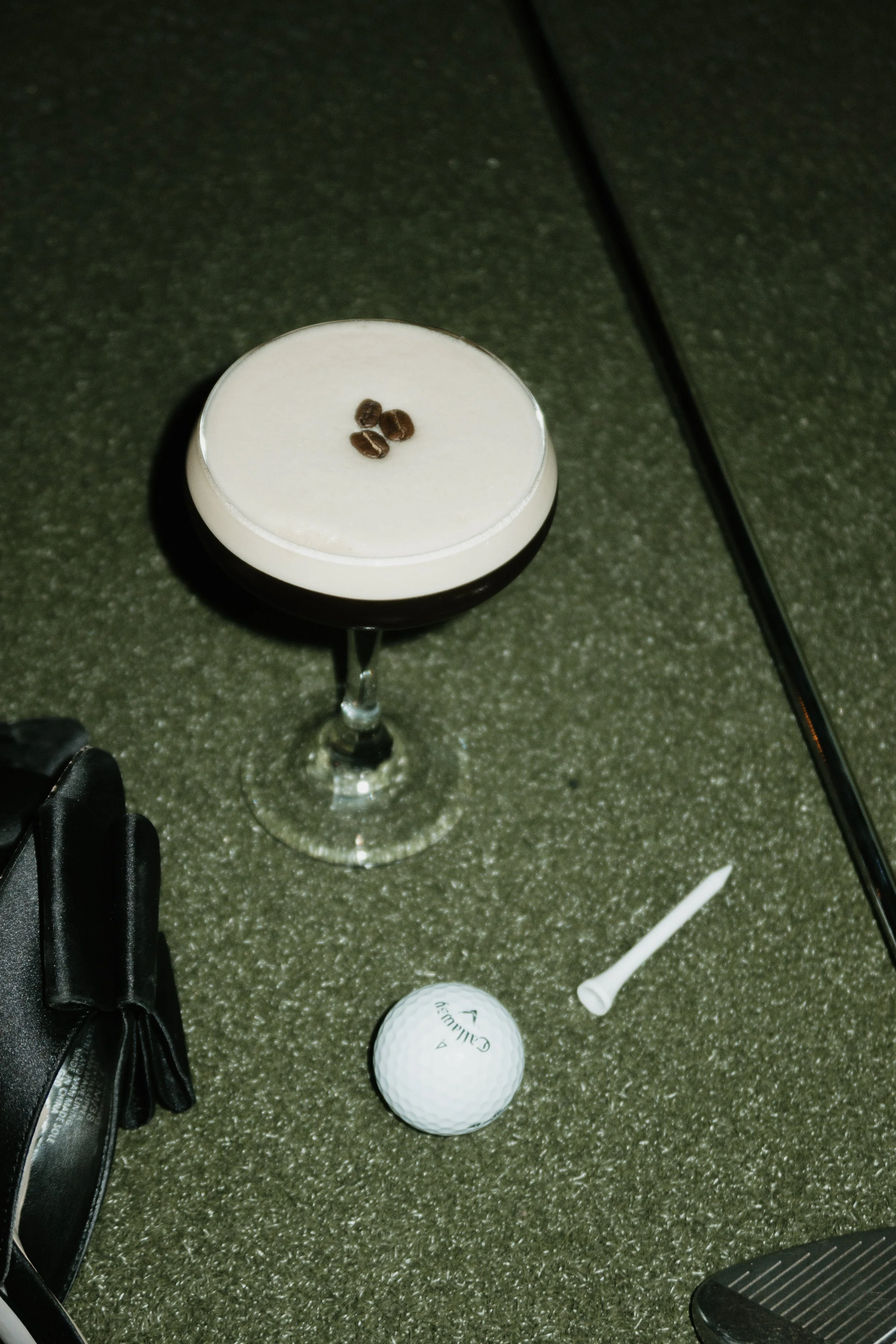 A tall glass of a white beverage with coffee beans on top, a golf ball, a white golf tee, and some golf club grips on a greenish indoor carpet.