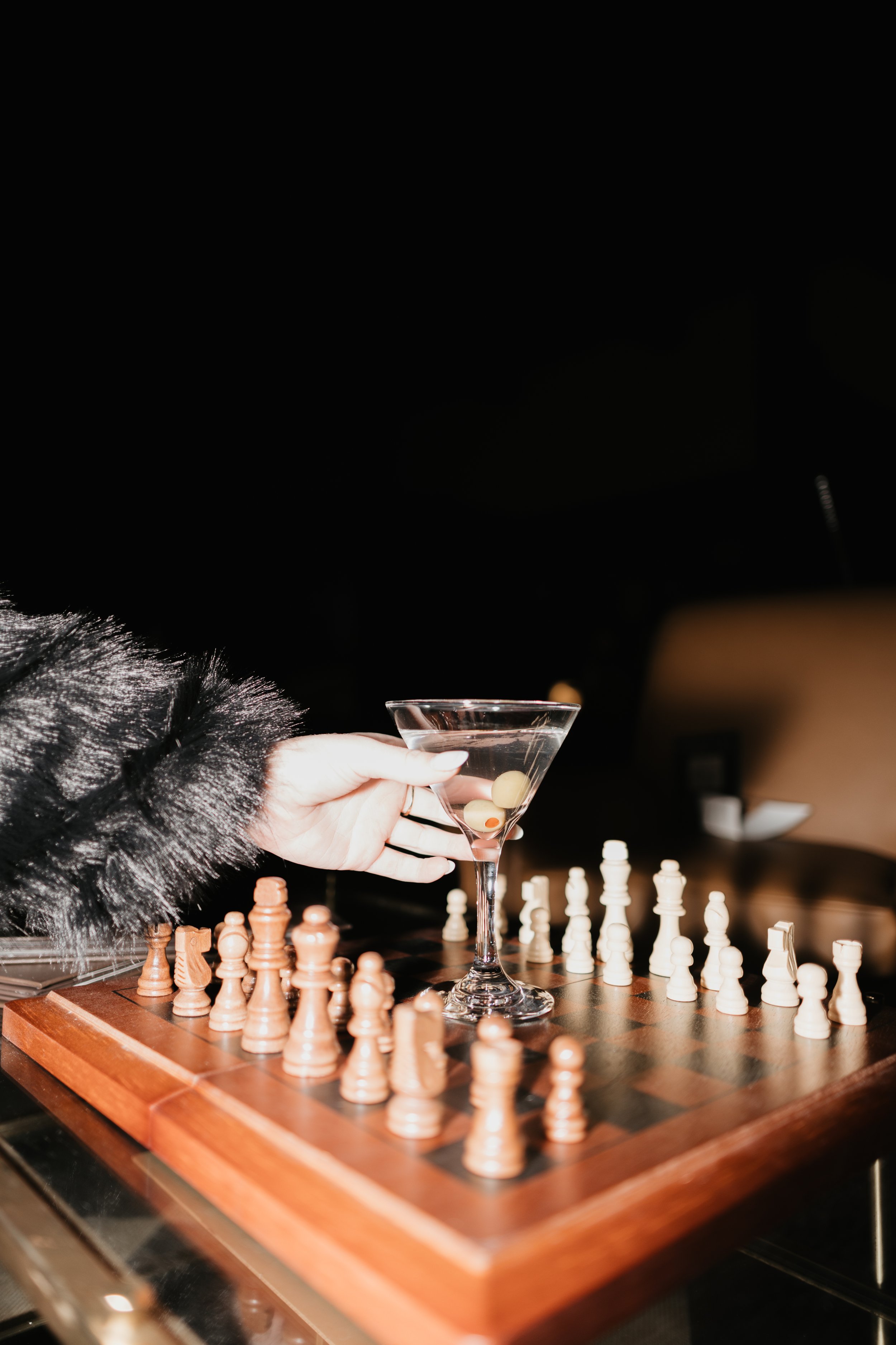 A person wearing a black fur coat playing chess in front of a chessboard, with a hand holding a martini glass with olives inside, on a dark background.