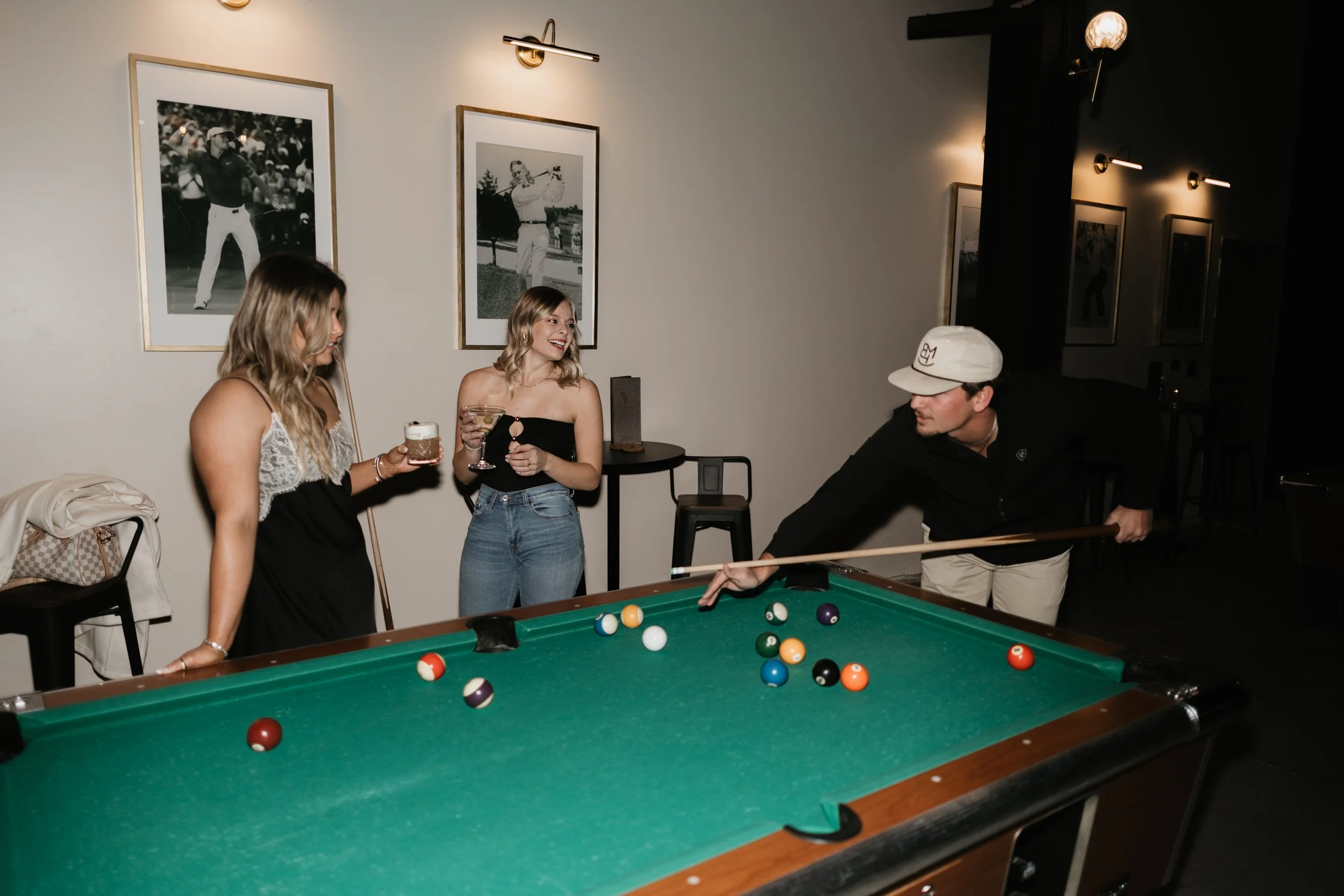 Three women and one man playing pool in a social setting, with two women holding drinks watching the game, and the man aiming with a cue stick.