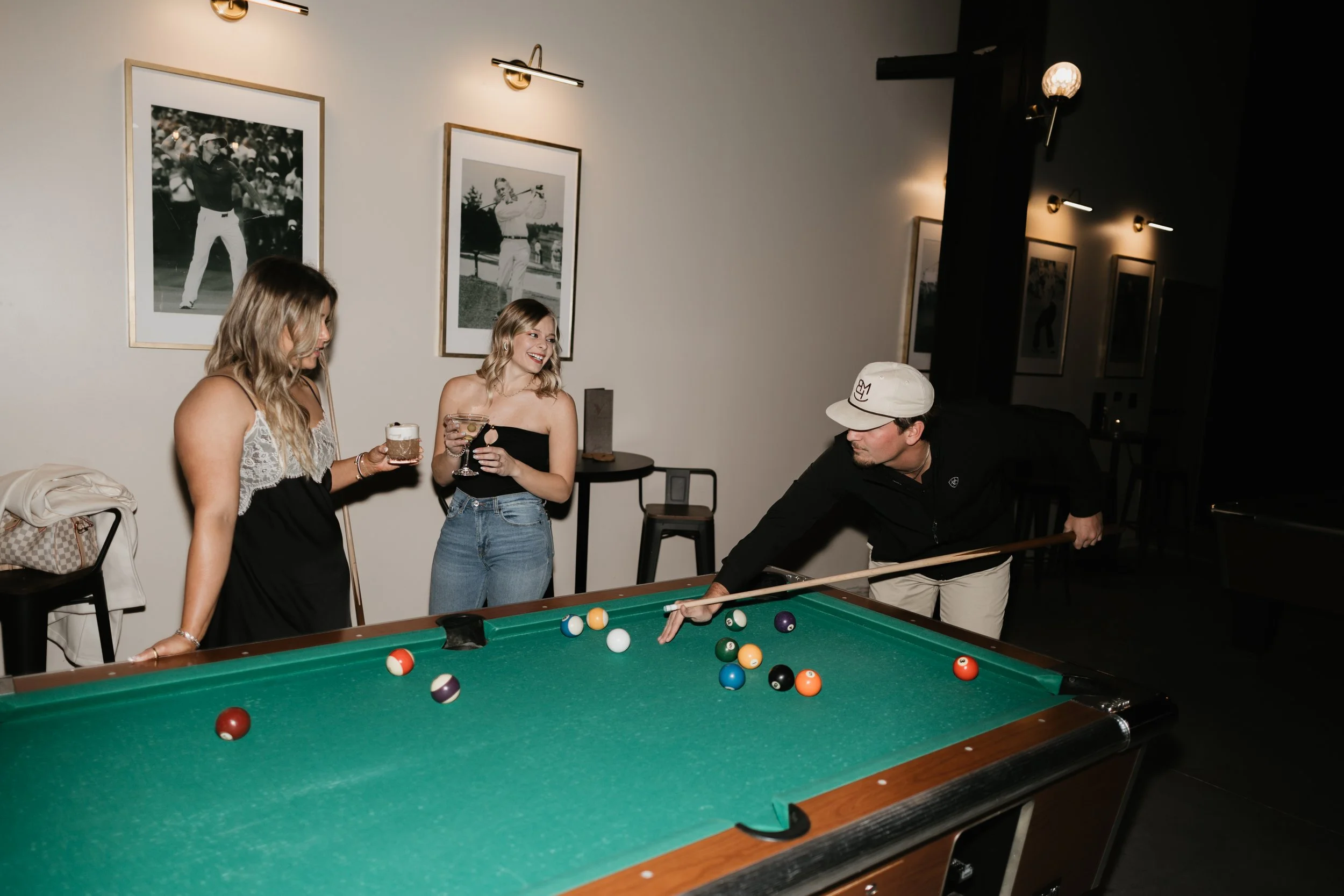 Three women and one man playing pool in a dimly lit room decorated with black and white framed photographs of golfers.