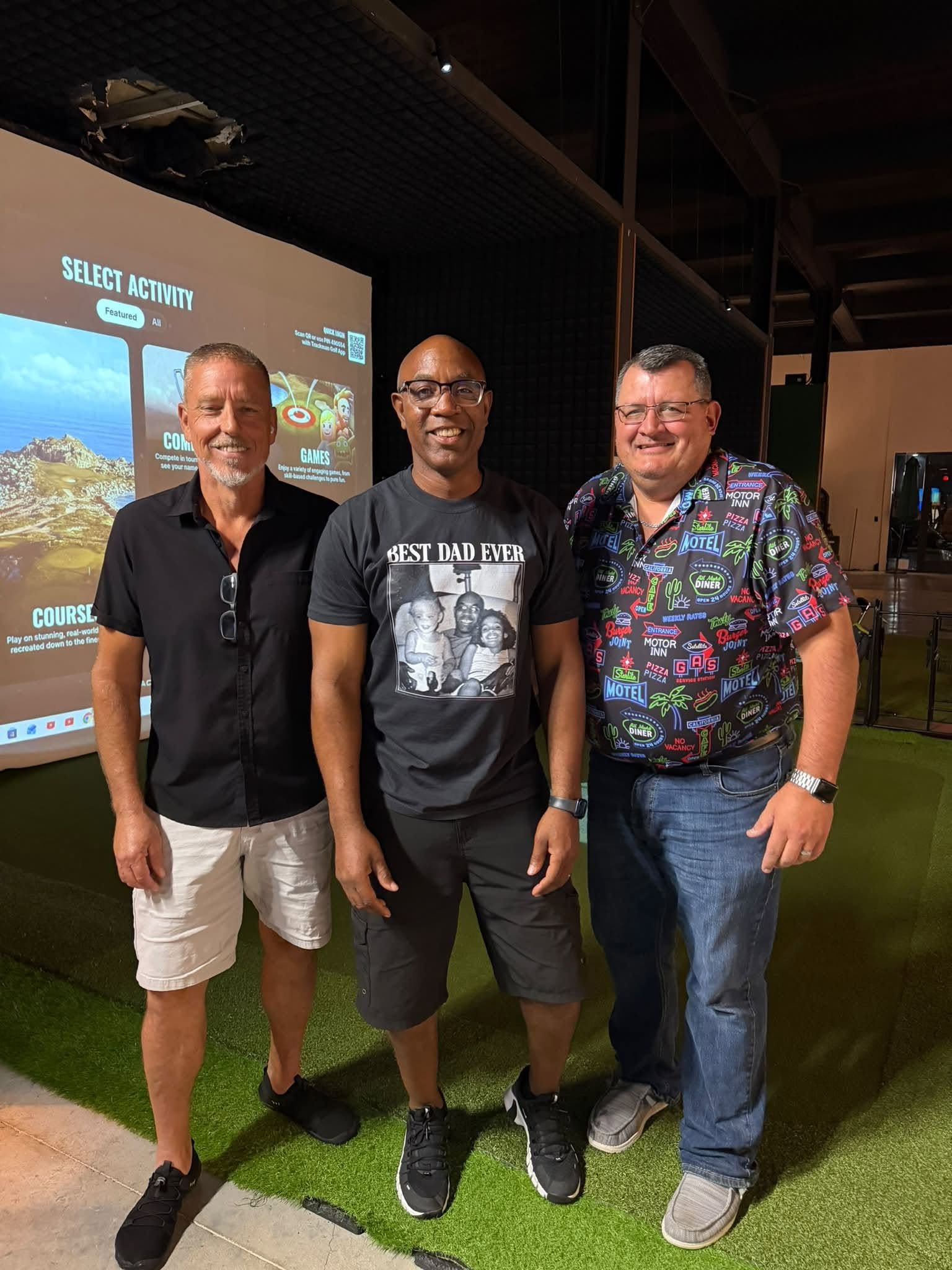 Three men standing together indoors, smiling at the camera. Behind them is a screen with the words 'Select Activity' and images of outdoor locations.