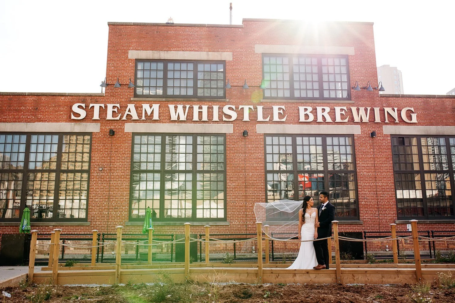 A bride and groom stand outside a red brick building labeled 'Steam Whistle Brewing,' with sunlight shining from above, holding a transparent umbrella.