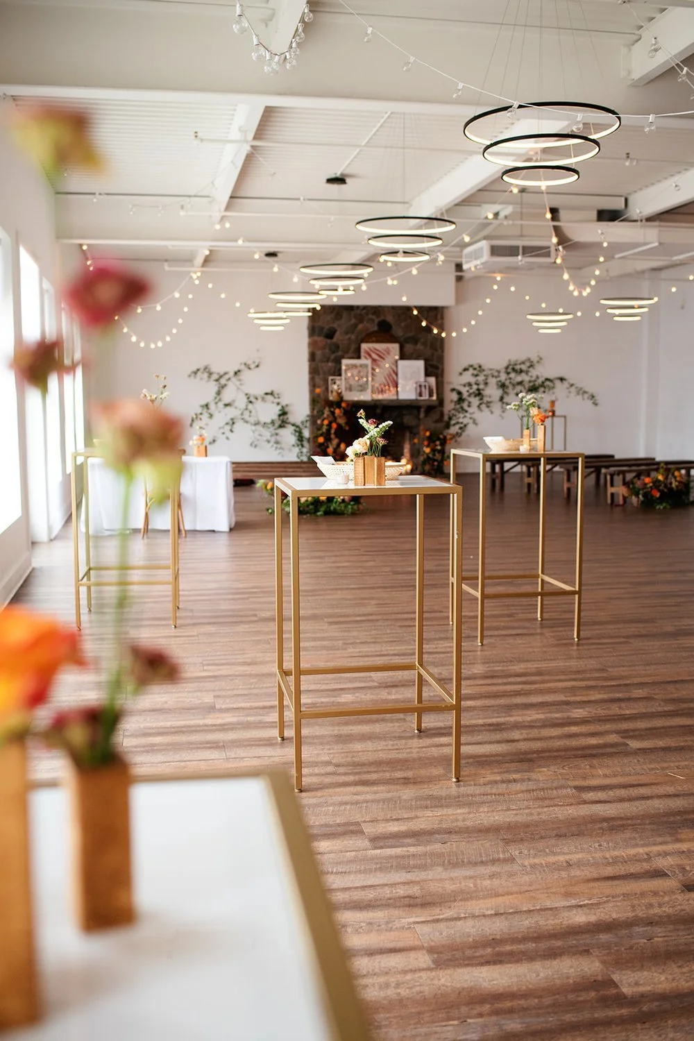 Empty event space with wooden floors, gold tables with flower arrangements, string lights, modern circular ceiling lights, and a stone fireplace decorated with flowers in the background.