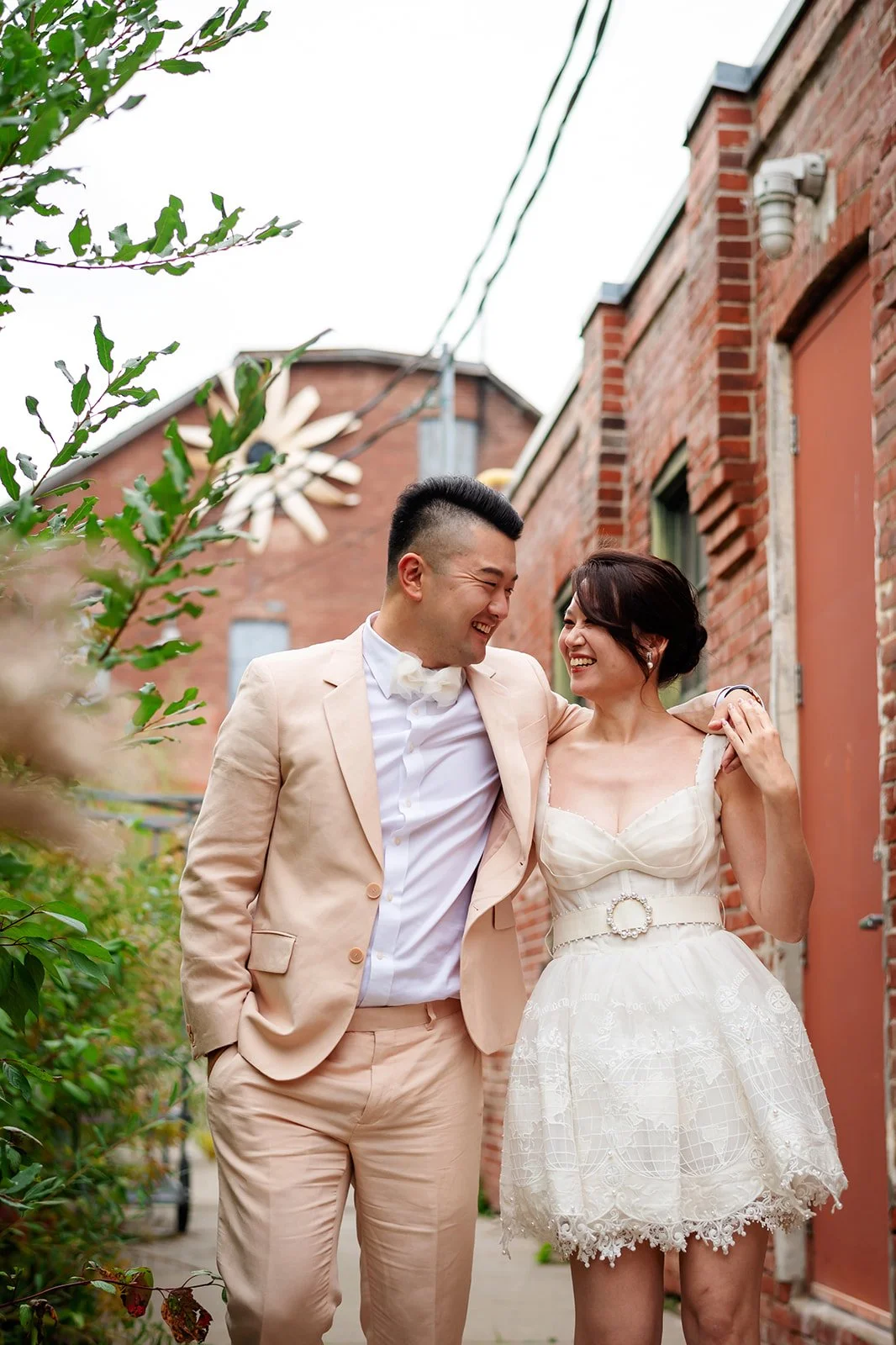 A happily smiling couple, dressed in wedding attire, walking arm-in-arm outdoors near red brick buildings and greenery.